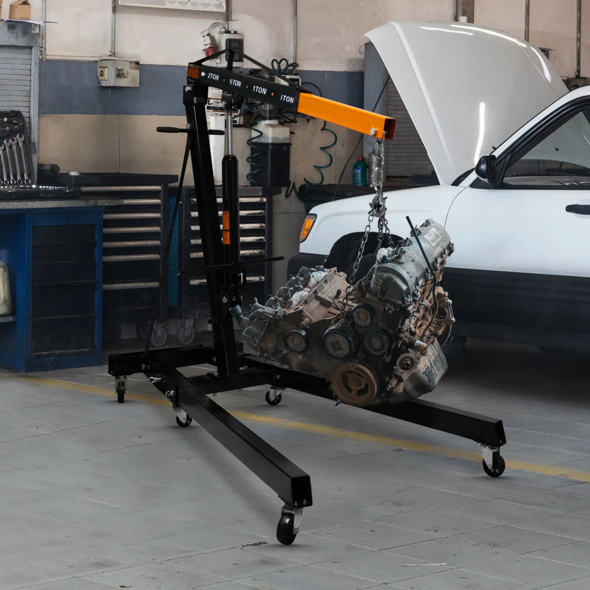 Hydraulic engine crane in black and orange, lifting a heavy engine in a garage setting, showcasing robust wheels and adjustable height