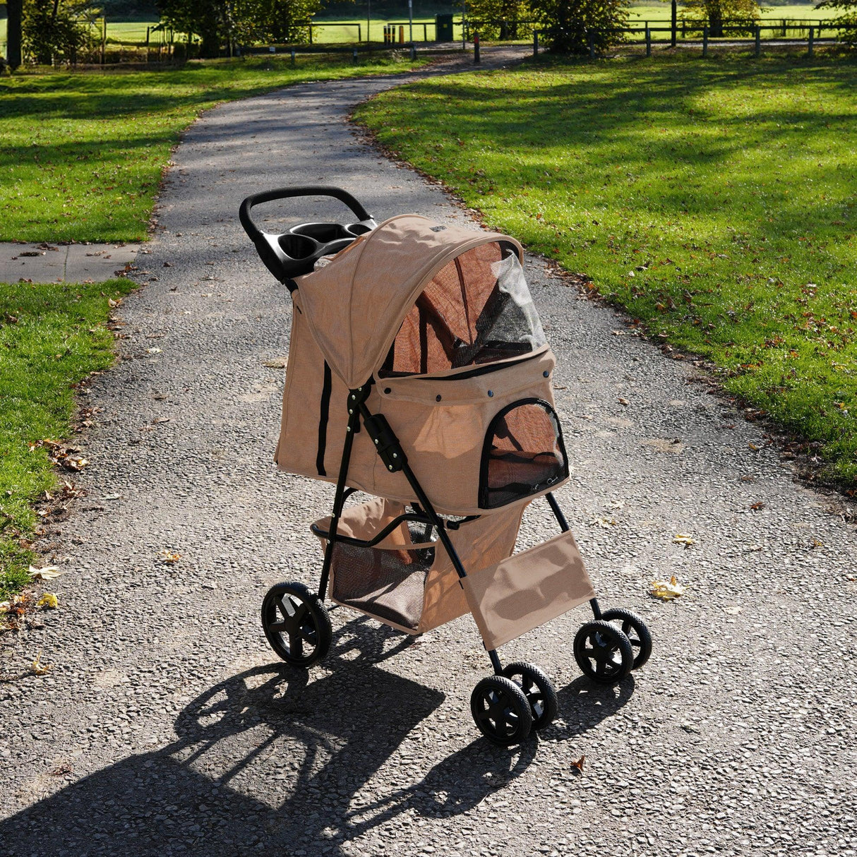 Woven beige pet stroller with rain cover, featuring a mesh canopy, cup holders, and sturdy wheels, set on a gravel path in a park