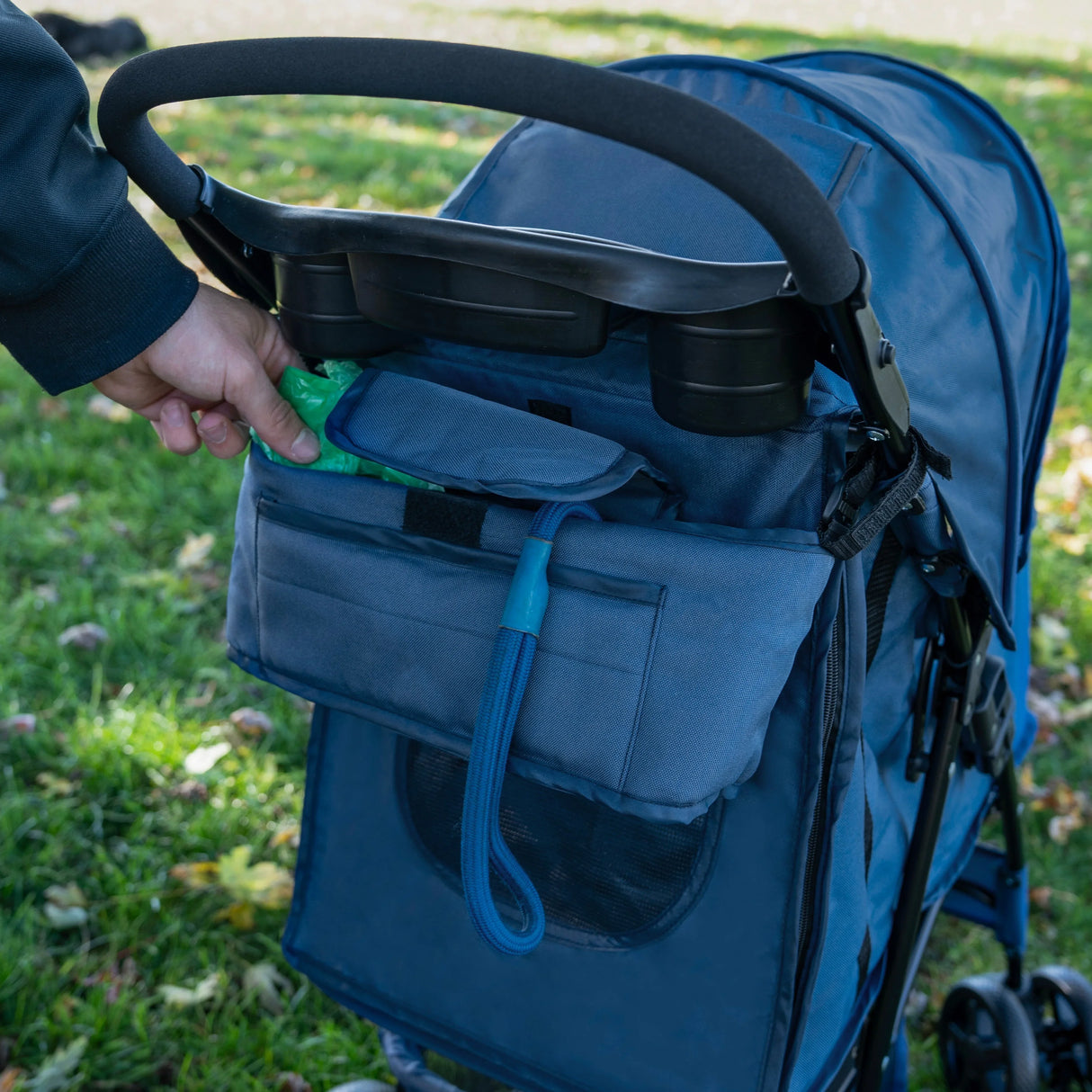 Navy blue pet stroller caddy bag with multiple pockets, hand reaching for a green waste bag, set against a grassy background