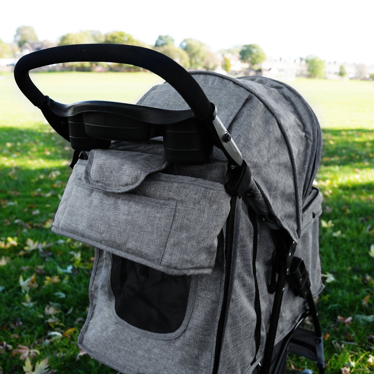 Woven grey pet stroller caddy bag featuring a spacious pocket, mesh window, and sturdy handle, set against a grassy park background
