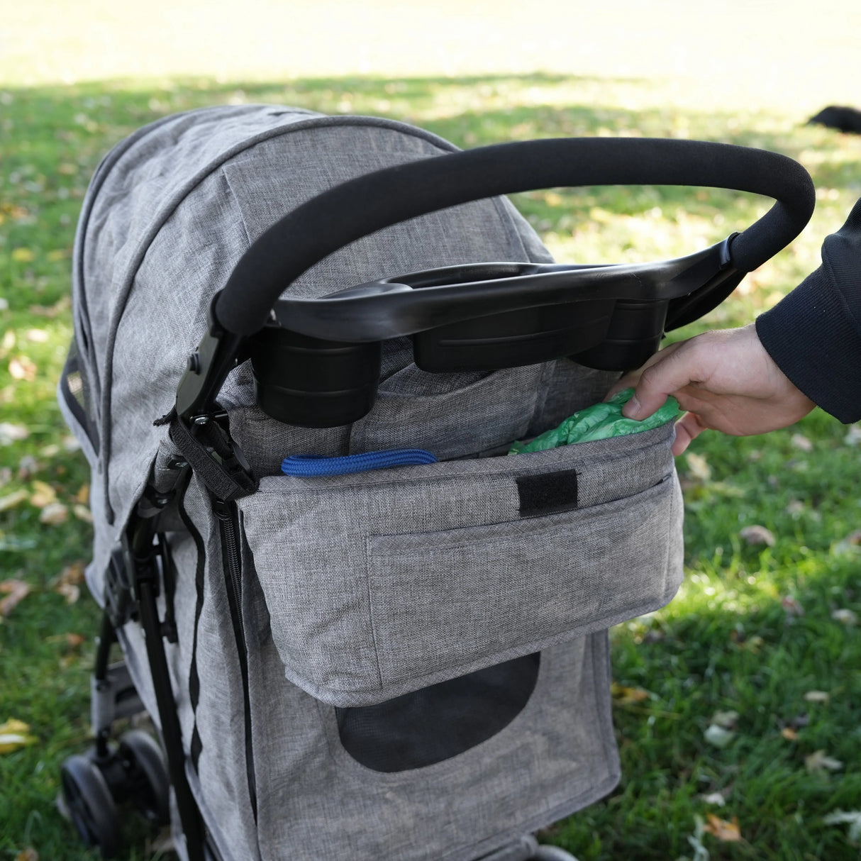 Woven grey pet stroller caddy bag with multiple pockets, holding essentials, attached to a stroller in a park setting