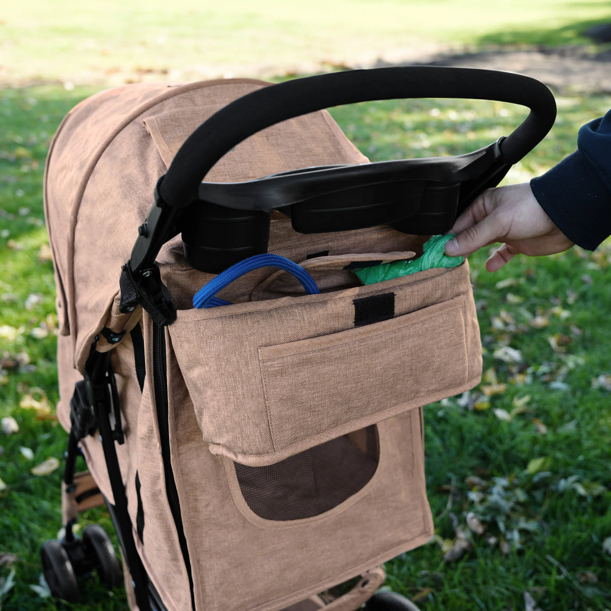 Woven beige pet stroller caddy bag with front pocket, holding dog waste bags, attached to a stroller handle in a grassy park setting