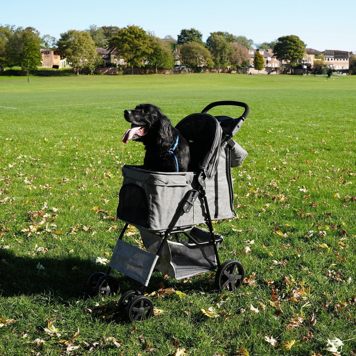 Pet stroller in woven grey with a rain cover, featuring a spacious caddy bag, parked on a grassy field with a happy dog inside