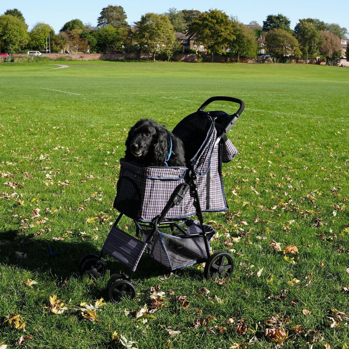Pet stroller in blue tartan with a rain cover, featuring a caddy bag, parked on a grassy field with a dog inside