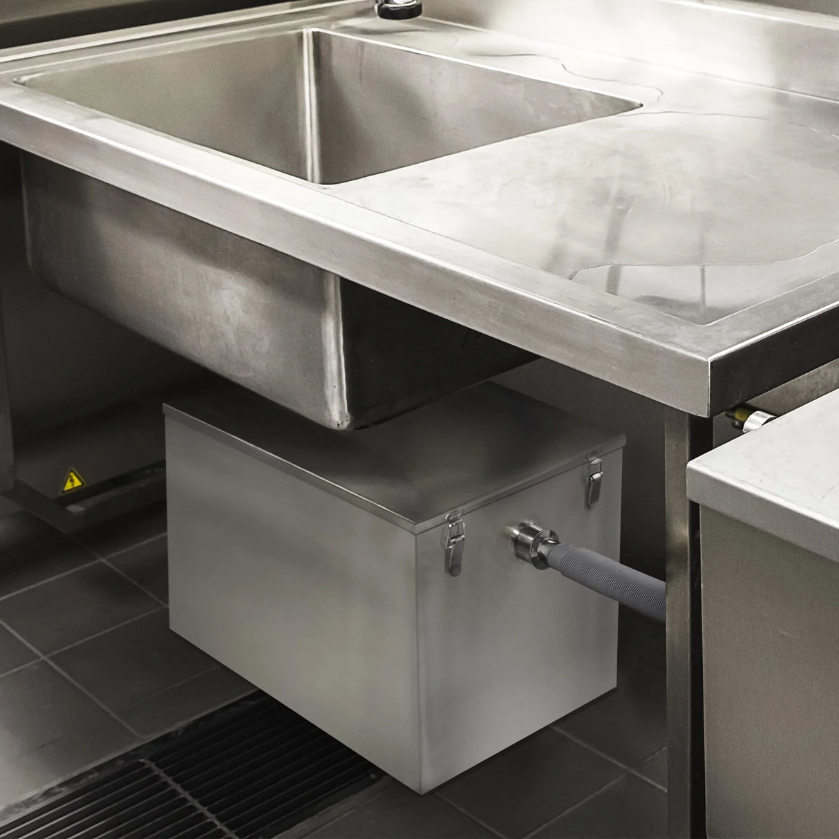 Stainless steel KuKoo grease trap, 96 litres, installed under a commercial kitchen sink with a grey drainage hose attached