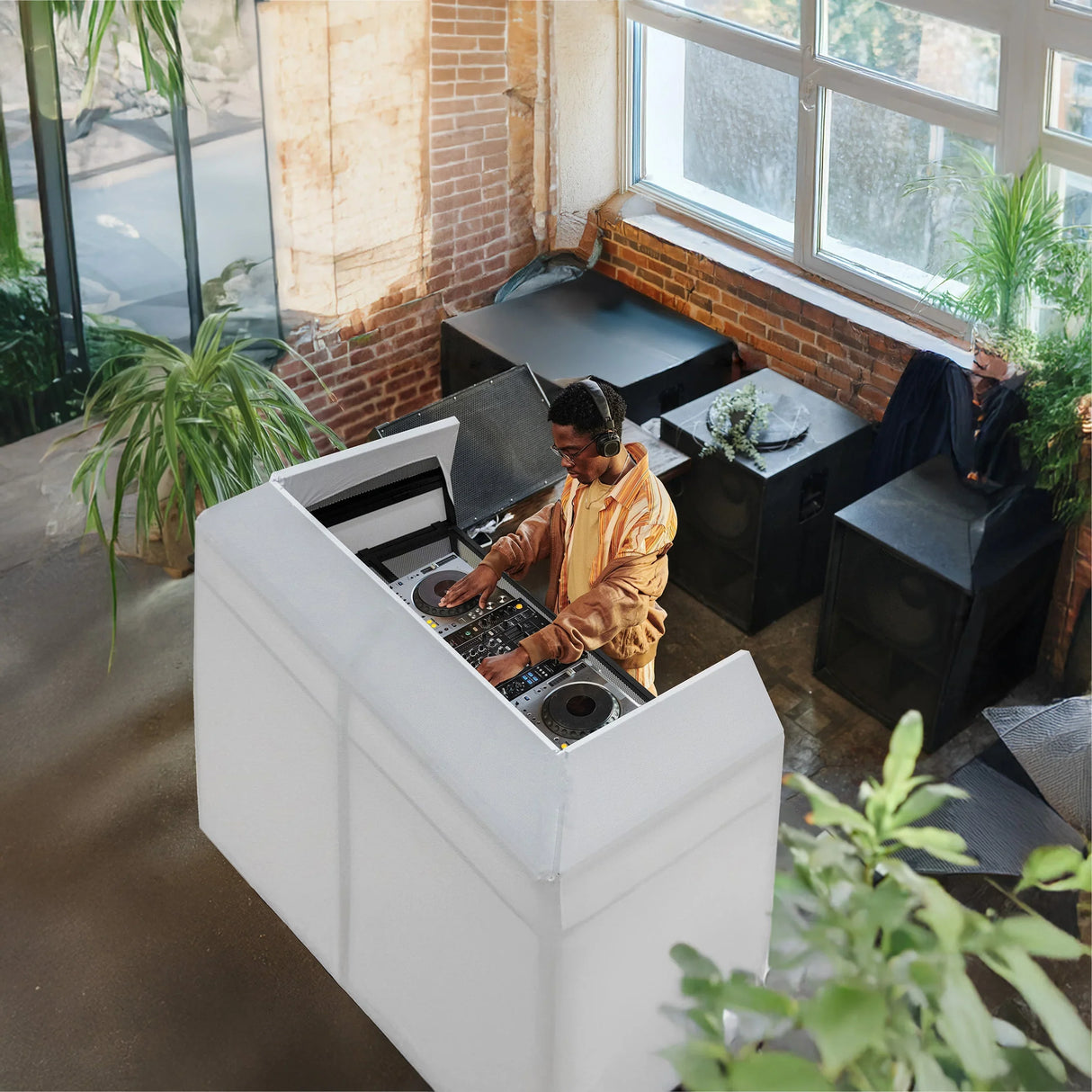 DJ Disco Booth Pro in a bright venue, featuring a sleek white design, DJ mixing music, surrounded by plants and speakers