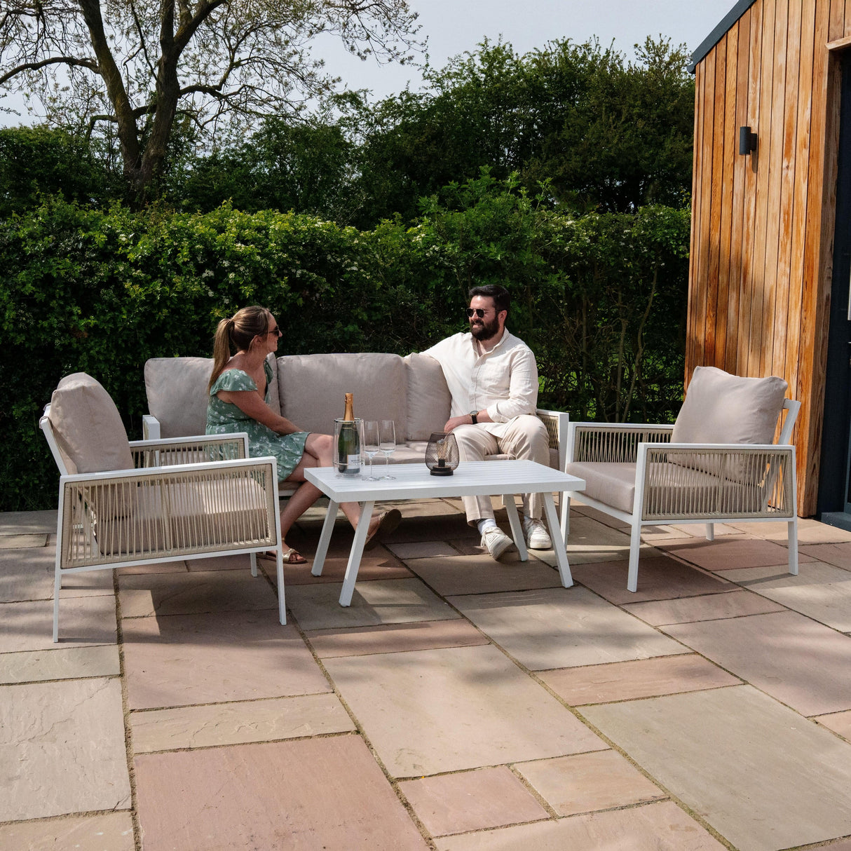 Aluminium 4 piece sofa set in white with plush beige cushions, arranged on a patio, featuring a coffee table and greenery background