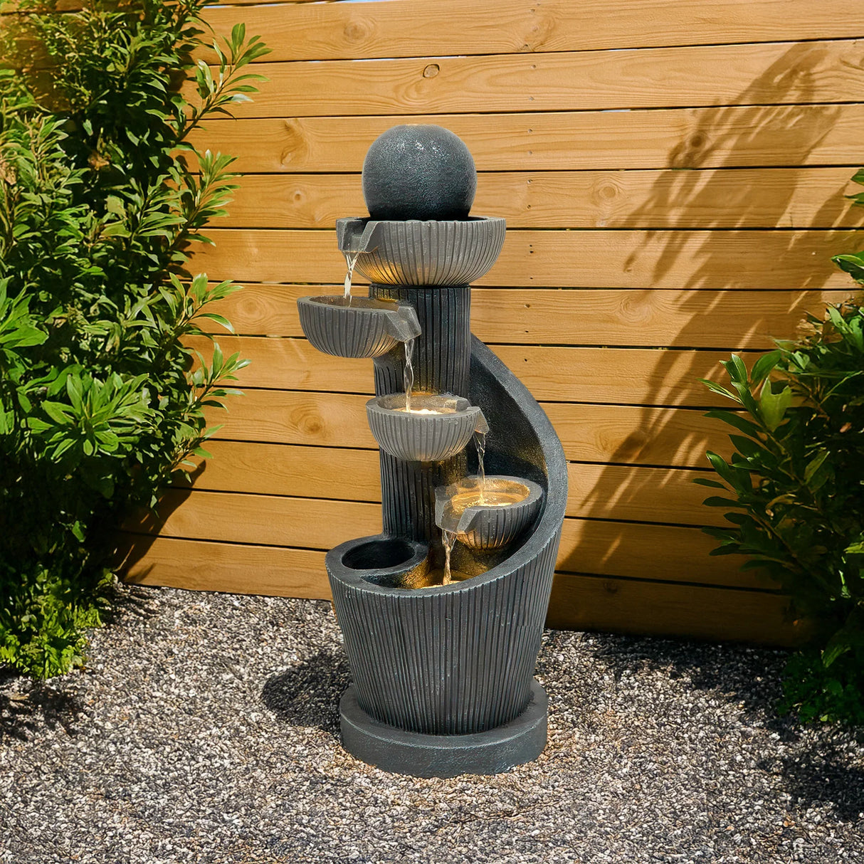 Elegant electric water feature with spiral springs, featuring cascading water from three bowls, set against a wooden backdrop and lush greenery