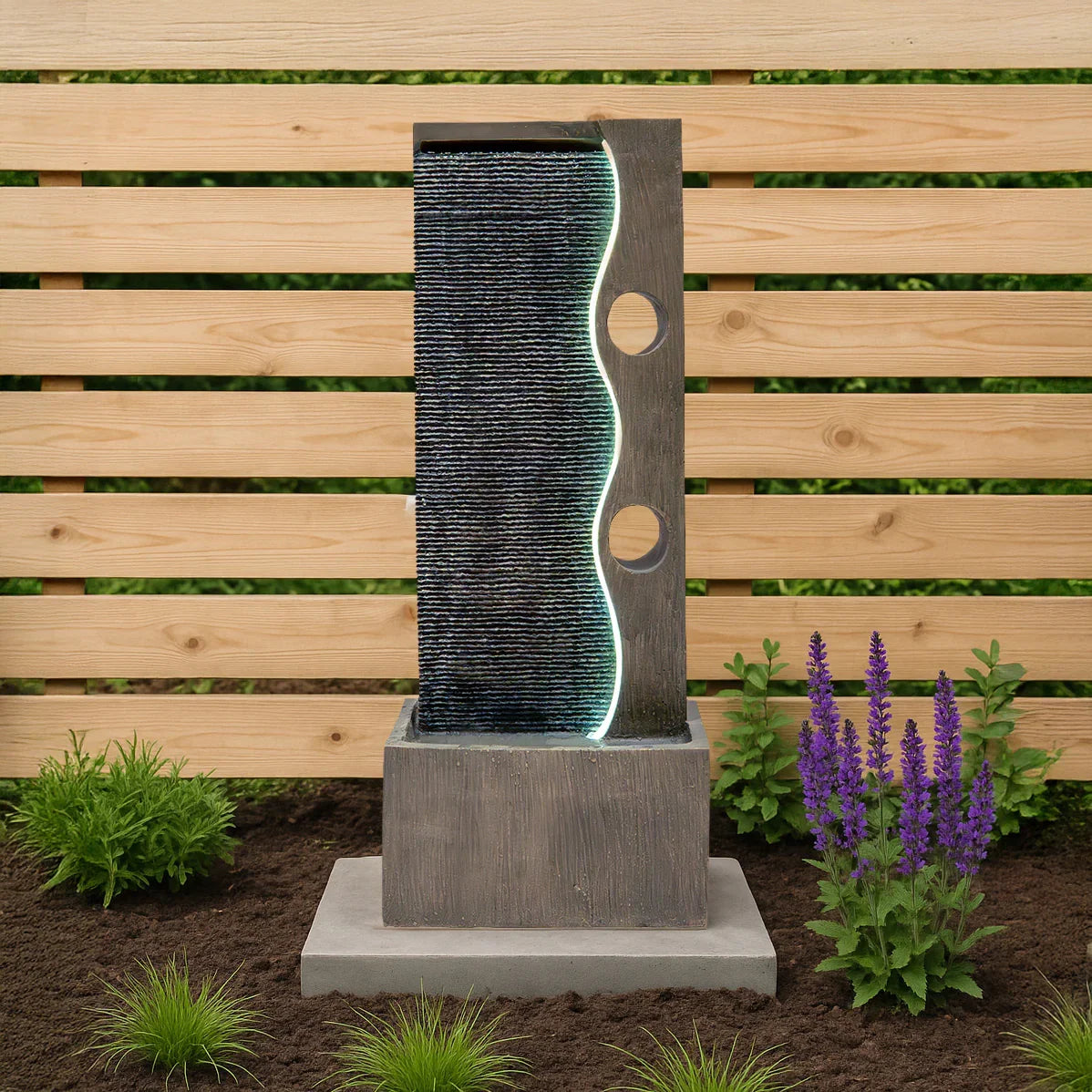 Electric water feature Harmony Falls with a sleek gray stone design, illuminated flowing water, and lush greenery surrounding it