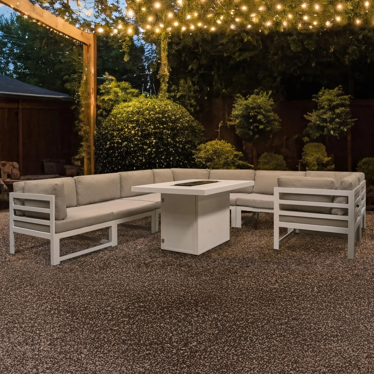 Modular garden furniture set in white with plush beige cushions and a central fire pit, surrounded by greenery and twinkling lights