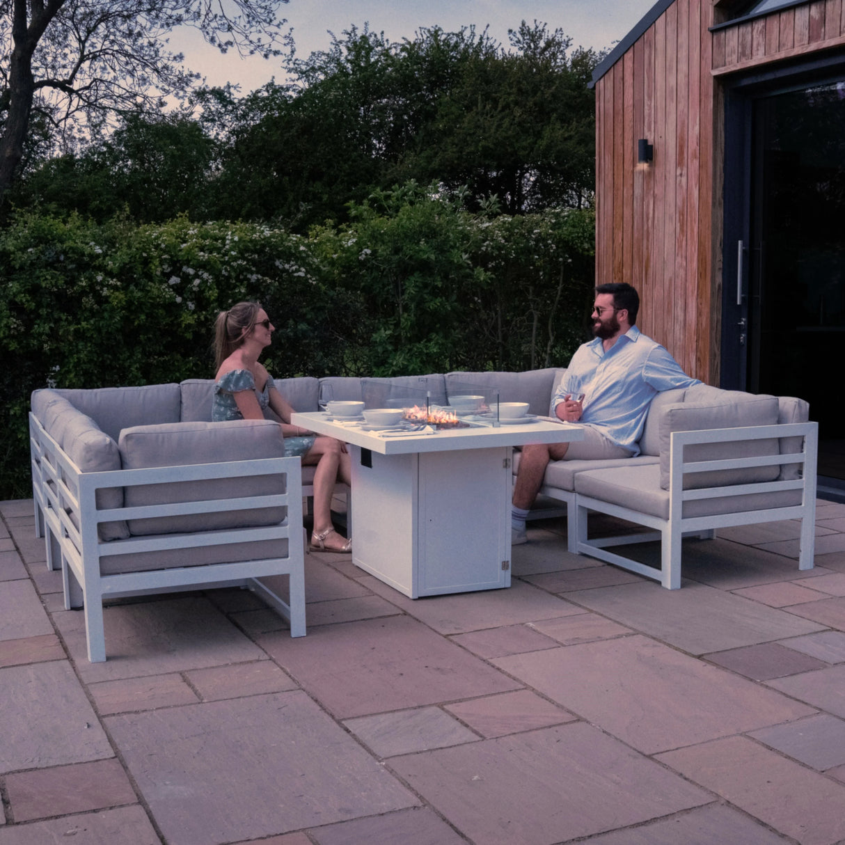 Modular white garden furniture set with fire pit, featuring soft cushions, arranged around a central table in a lush outdoor setting