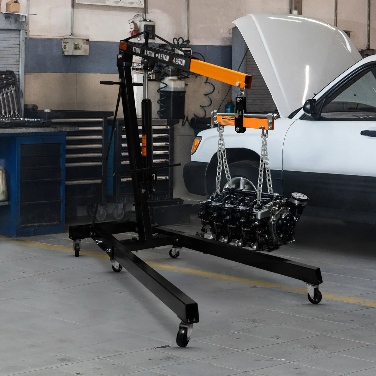 Heavy-duty engine load leveller with a black frame and orange accents, lifting an engine with chains in a garage setting