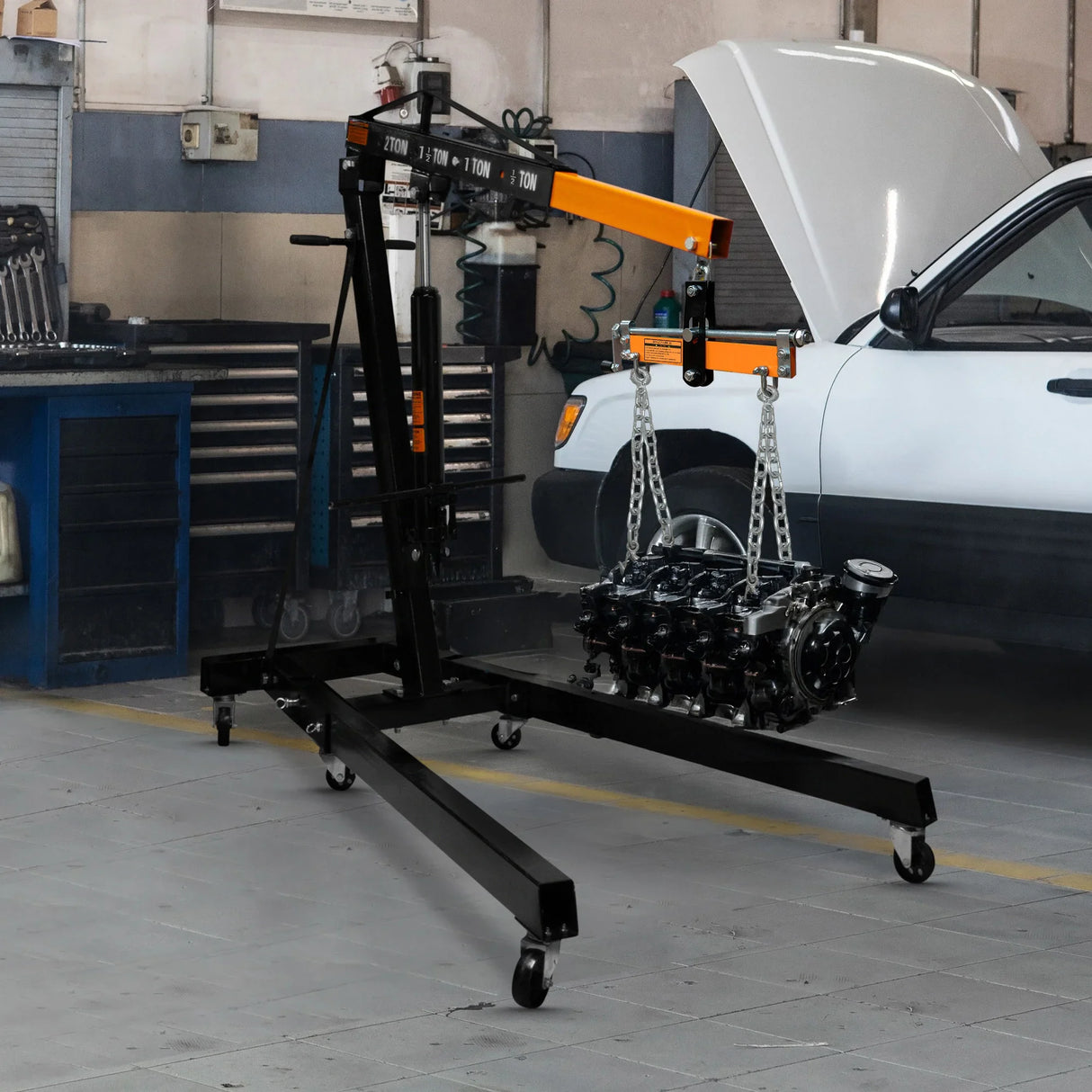 Engine crane with a 2-tonne capacity, featuring a sturdy black frame, orange load leveller, and an engine suspended by chains in a garage setting