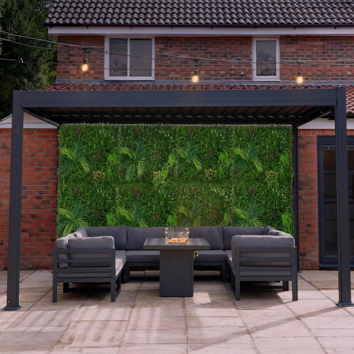Lush green Alina artificial wall tile backdrop enhances a modern outdoor seating area with a sleek black sofa and fire table