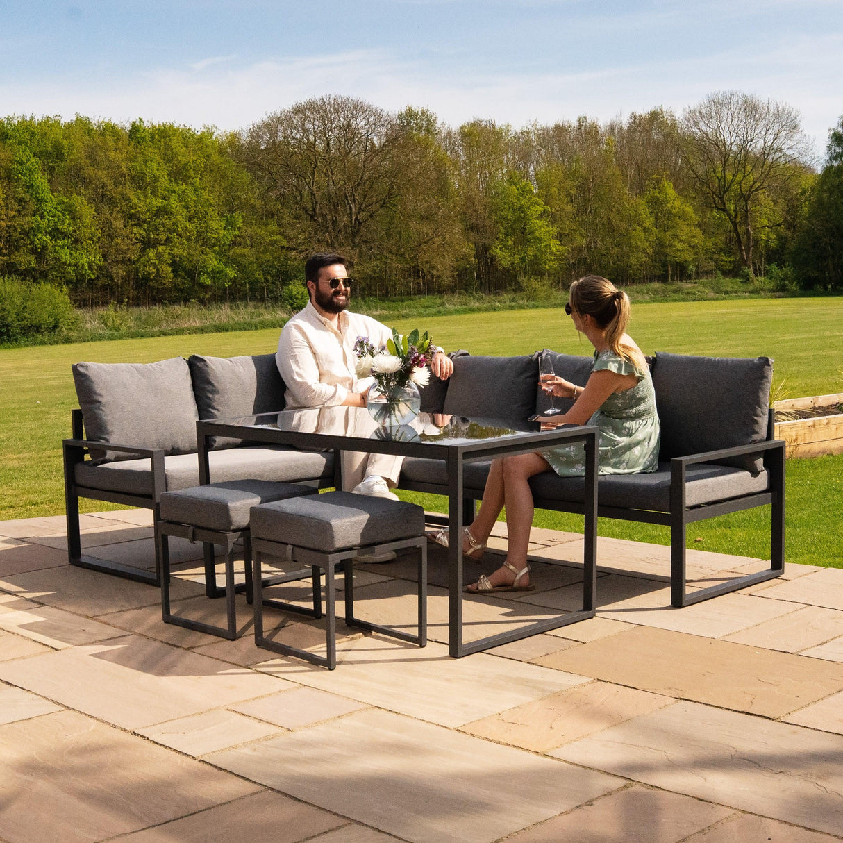 Anthracite grey aluminium corner sofa set with plush cushions, stylish glass table, and two matching stools in a sunny outdoor setting