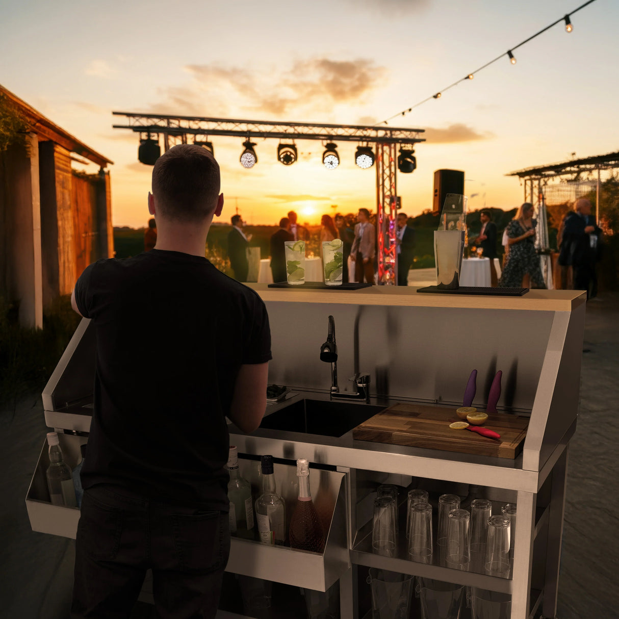 Mobile cocktail bar station with sleek stainless steel finish, wooden countertop, and vibrant cocktail ingredients, set against a sunset event backdrop