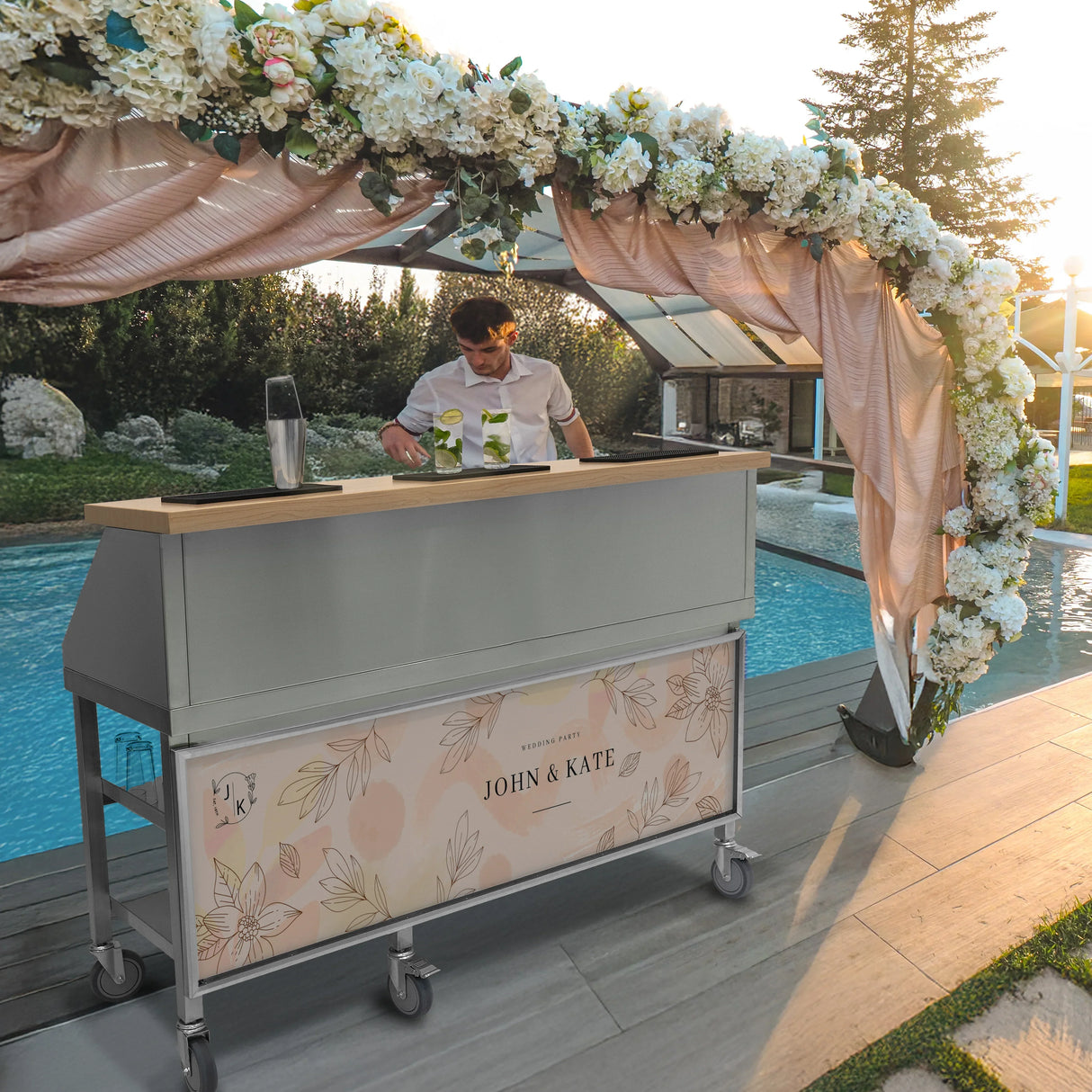Mobile cocktail bar station with a sleek gray design, wooden countertop, and personalized floral branding, set by a poolside event