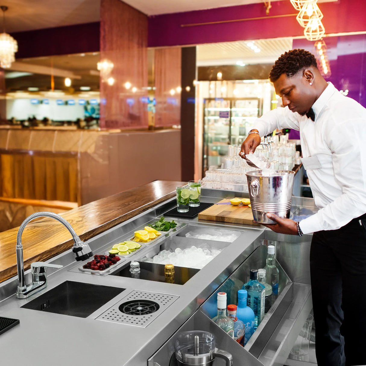 Modular cocktail bar corner unit with sleek stainless steel finish, ice compartment, and fresh garnishes, set in a vibrant lounge