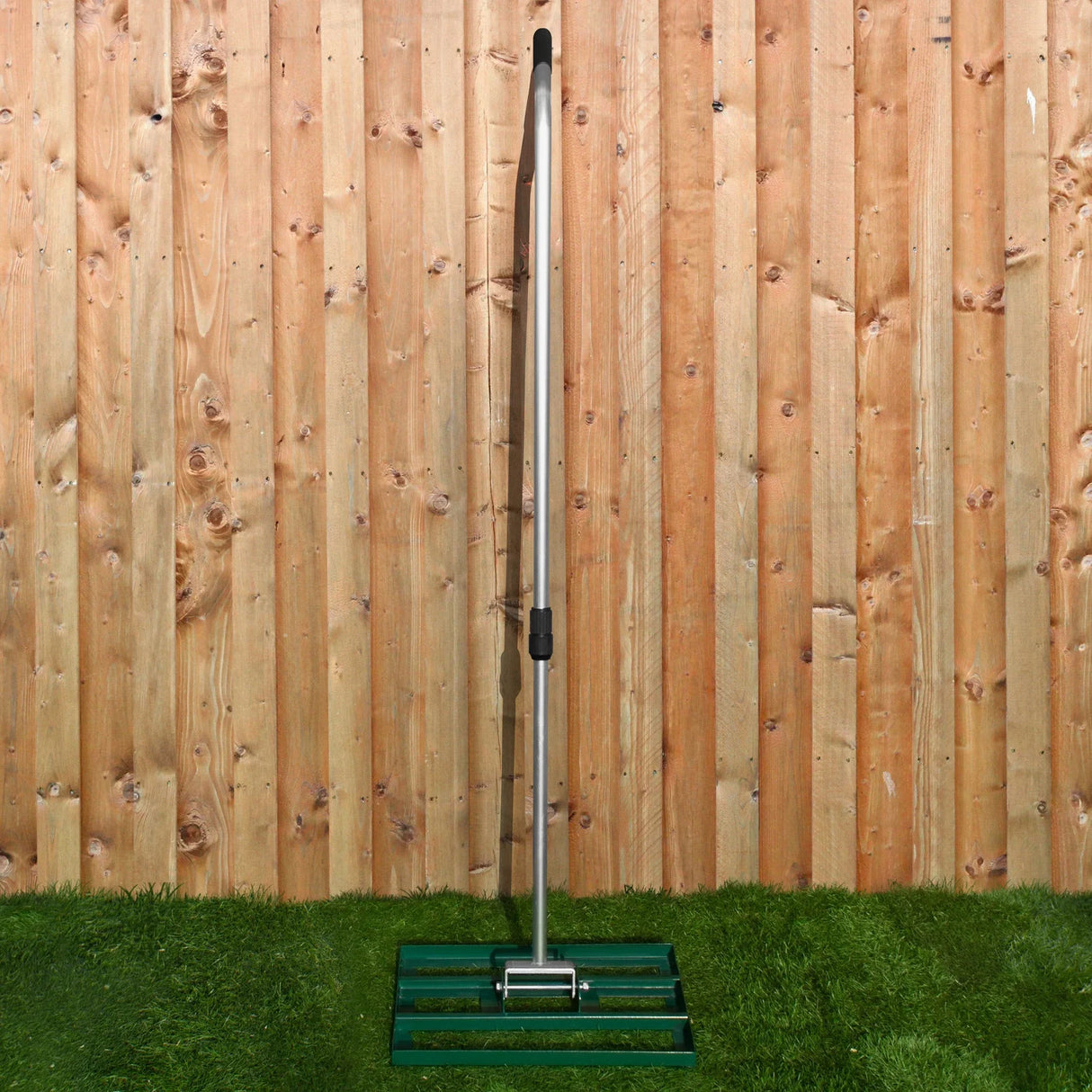 Green rotary spreader with a sturdy metal handle and a 50cm lawn leveller base, set against a wooden fence and grass background