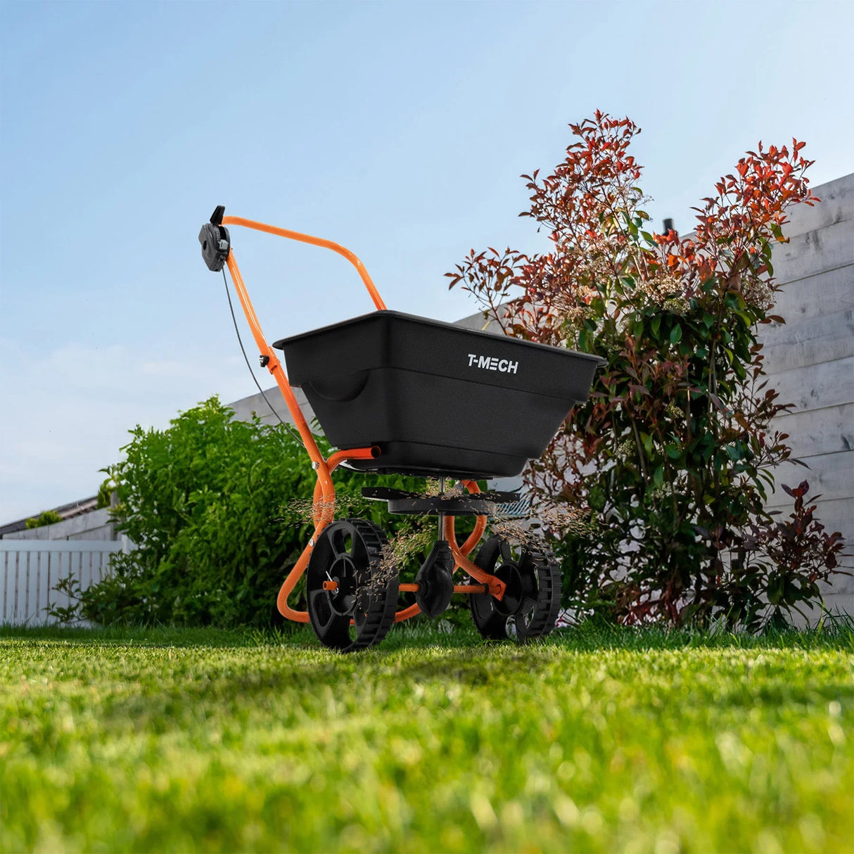 Rotary spreader with black hopper and orange frame on green grass, surrounded by plants and a wooden fence under a blue sky
