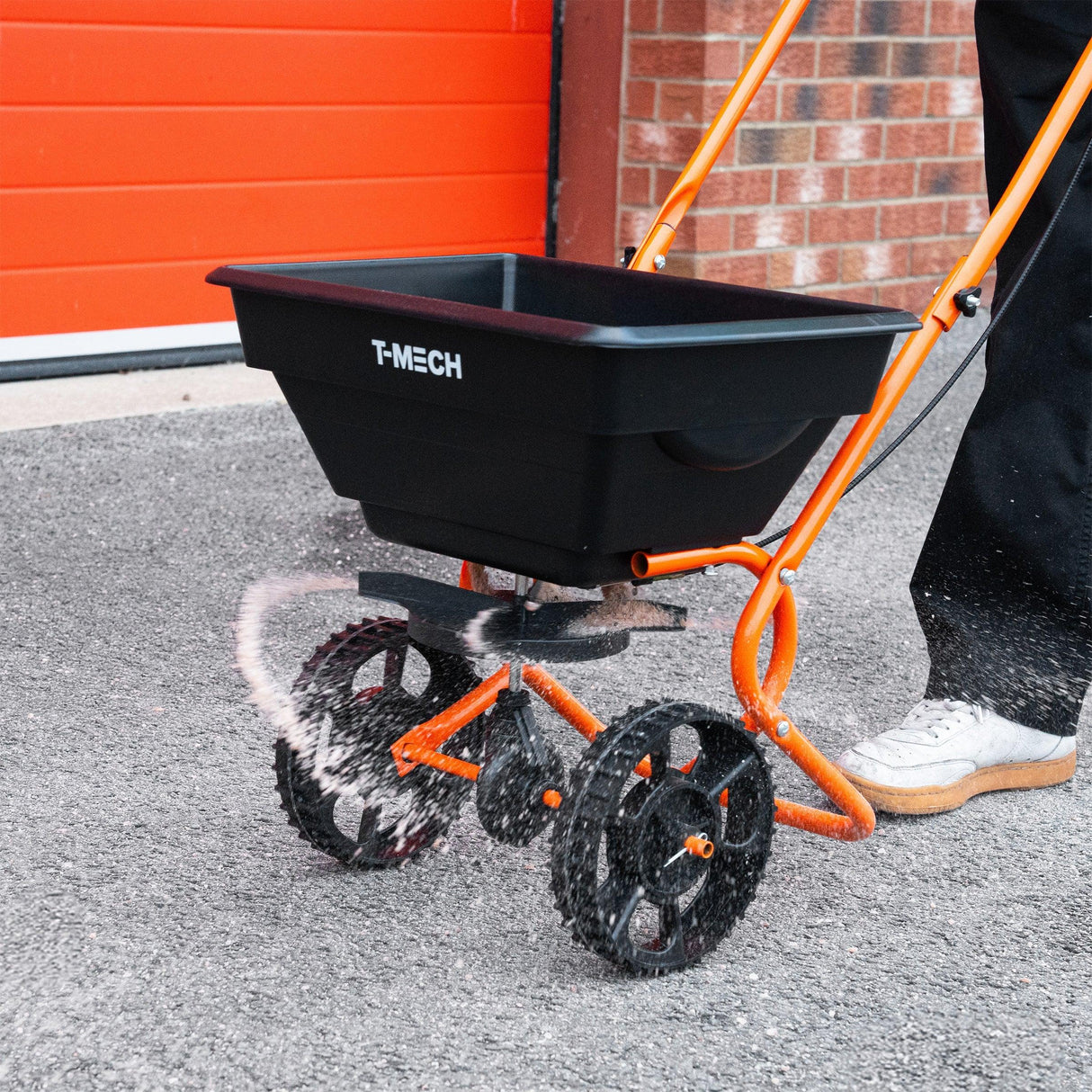 Rotary spreader with a black hopper and orange frame, featuring large wheels for easy maneuverability on various surfaces