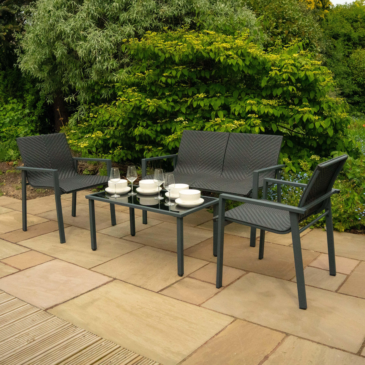 Black rattan patio furniture set featuring two armchairs and a glass-top coffee table, surrounded by lush greenery and stone paving