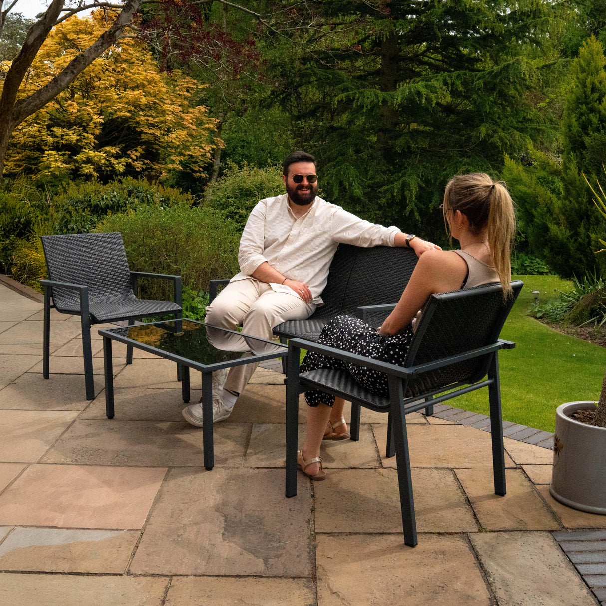 Black rattan patio furniture set featuring two chairs and a glass-top table, set in a lush garden with greenery and stone patio