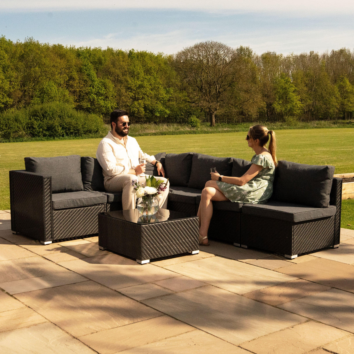 Rattan modular 7 piece furniture set in black, featuring plush gray cushions, arranged in a cozy outdoor seating area with greenery in the background