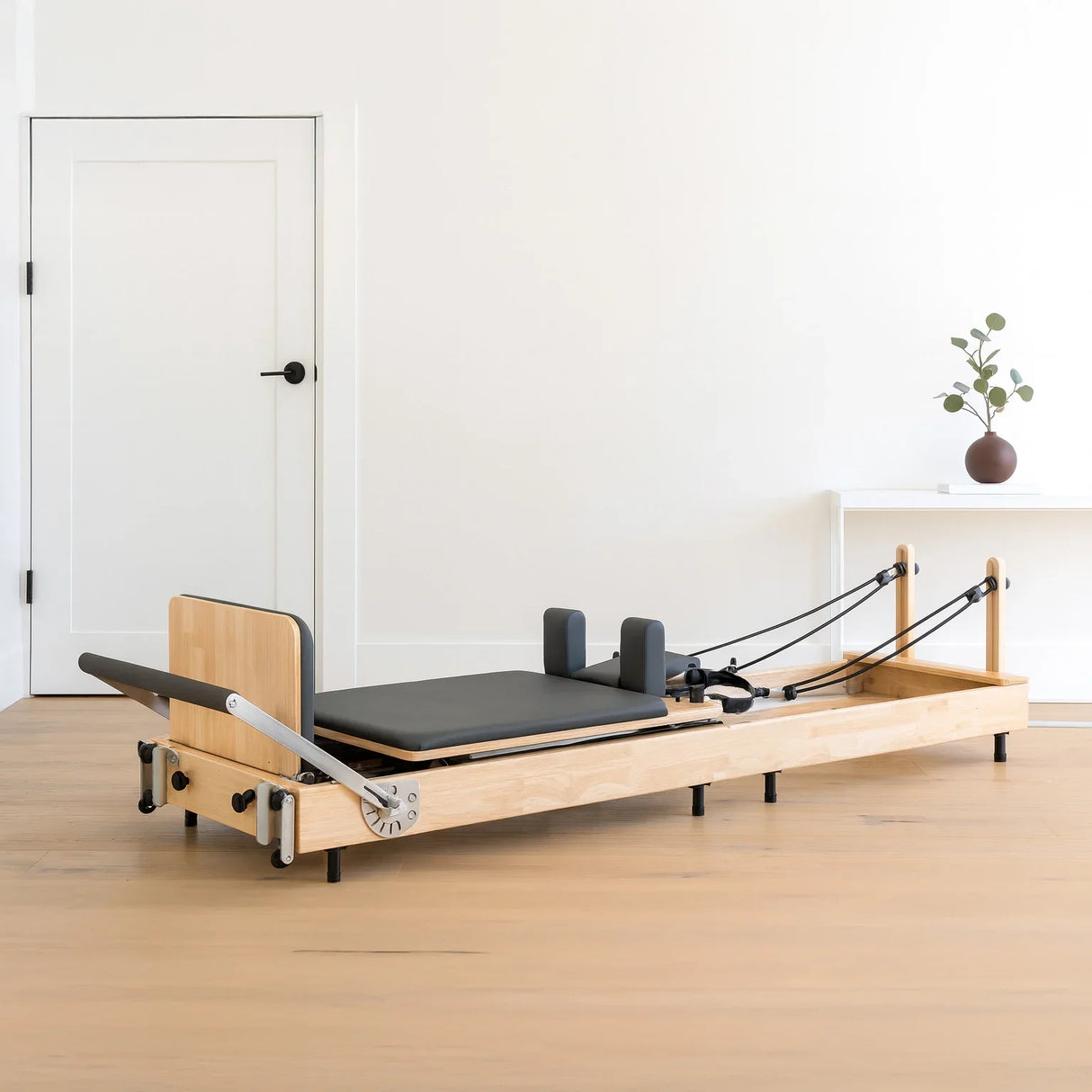 Pilates reformer machine in oak wood with a black padded platform, adjustable straps, and a minimalist design, set in a bright room