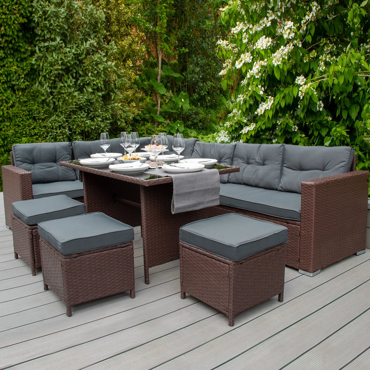 Rattan corner set with dark gray cushions, featuring a spacious table and matching ottomans, set in a lush garden environment