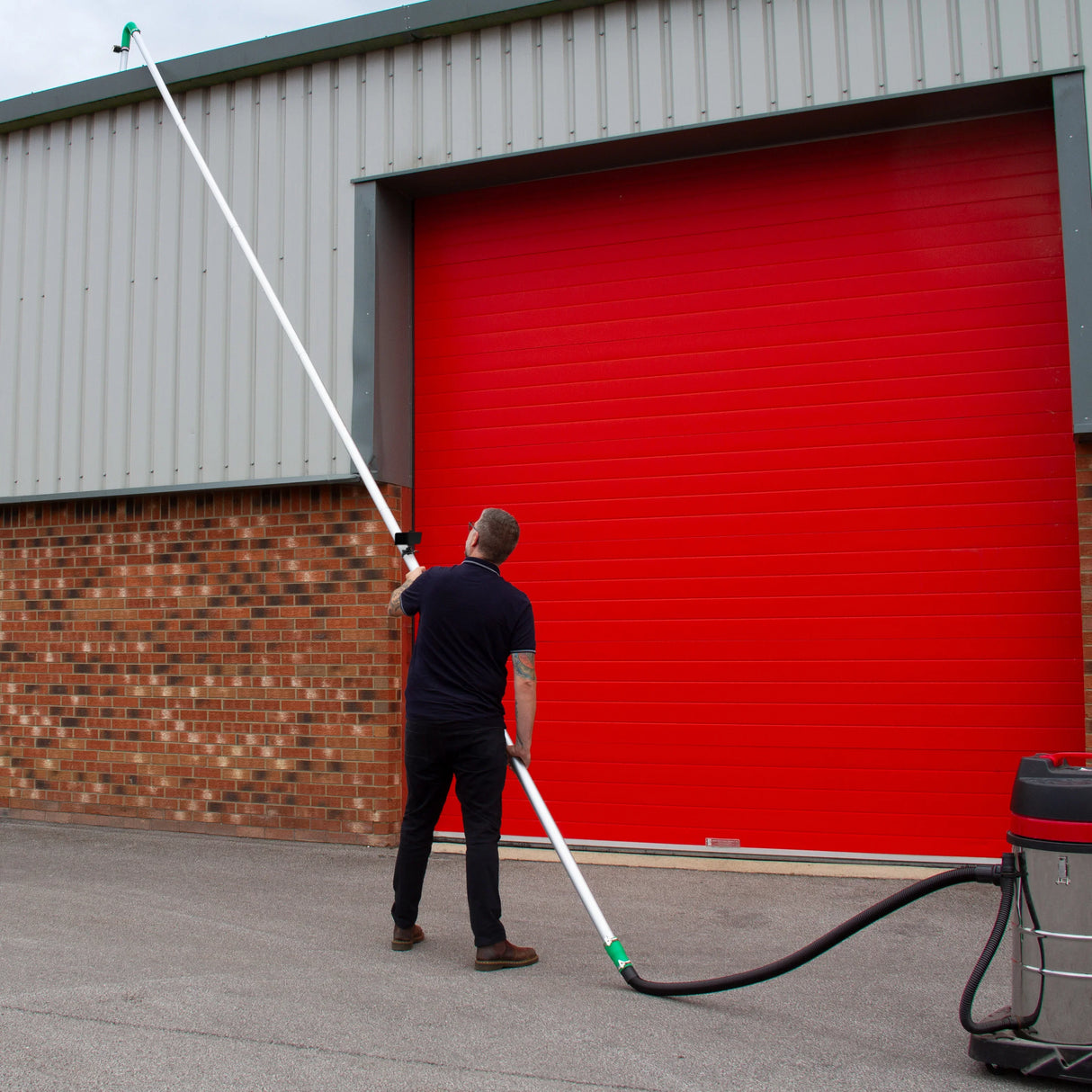 Gutter vacuum camera with a long pole, user aiming at a high gutter, red garage door in the background, outdoor setting