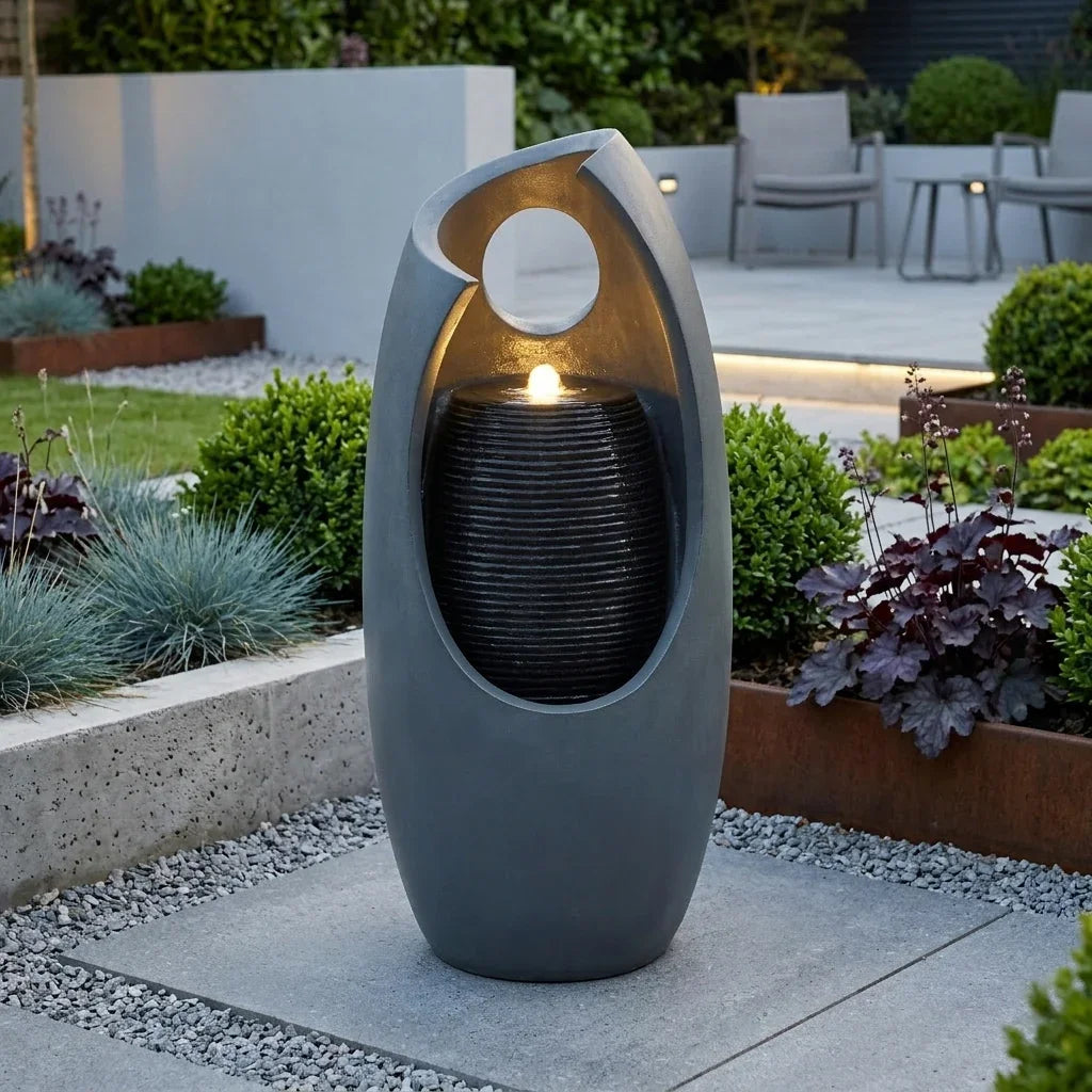 Sleek gray electric water feature with a modern design, featuring cascading water and a soft glowing light, surrounded by lush greenery