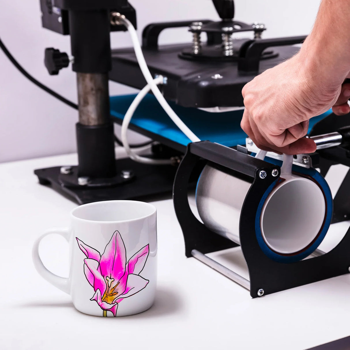 White ceramic mug with pink floral design next to a black and blue heat press machine on a white surface.