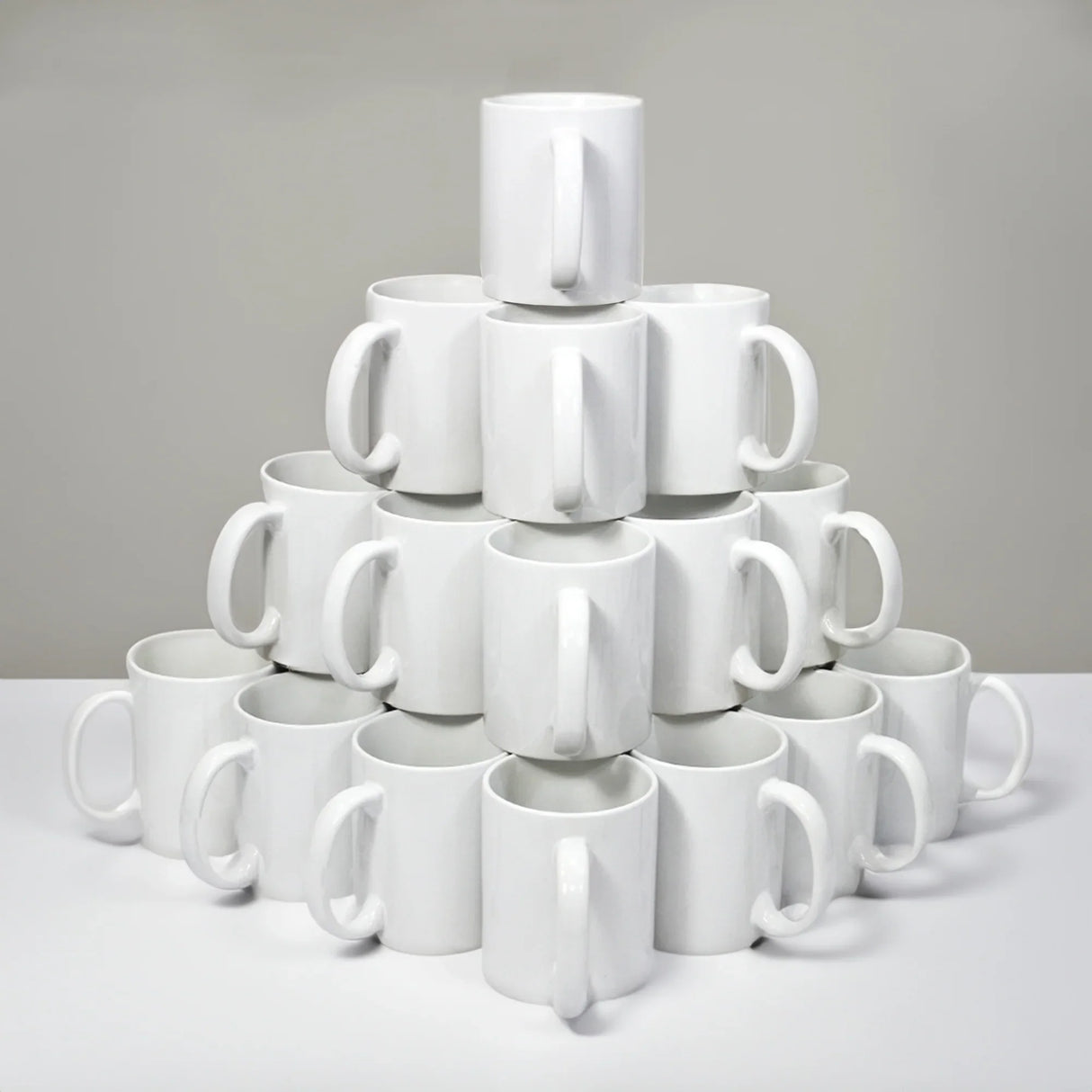 Stacked pyramid of plain white ceramic mugs with handles against a gray background.
