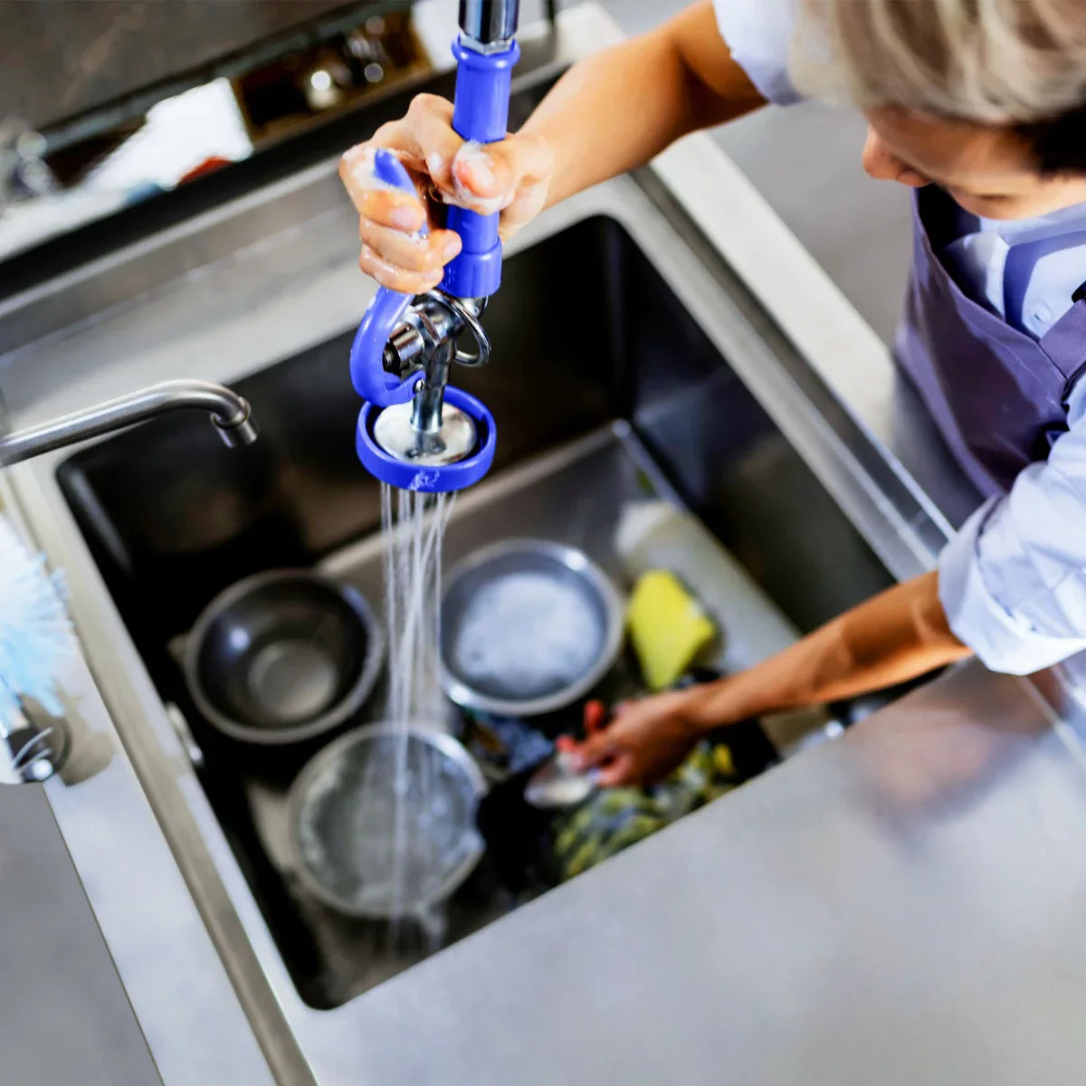 KuKoo Pre-Rinse Commercial Tap in use, featuring a blue handle, stainless steel sink, and water spraying over dirty dishes