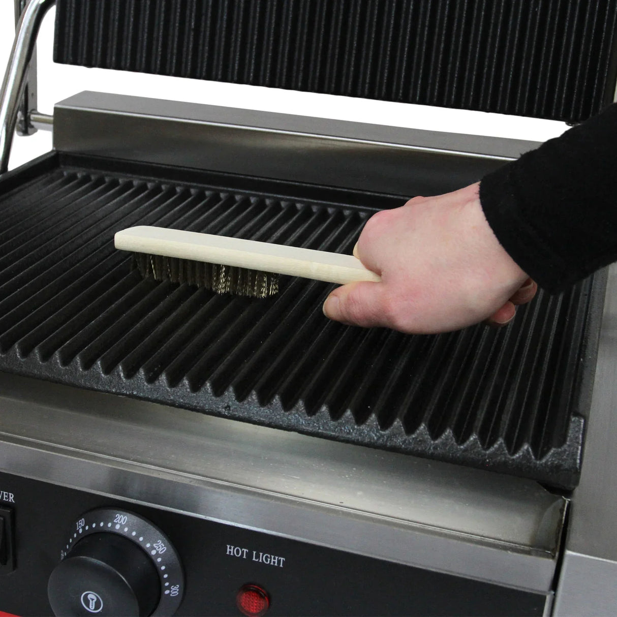 Grooved panini press with a hand using a wooden brush to clean the ridged grilling surface, showcasing its sturdy design and functionality