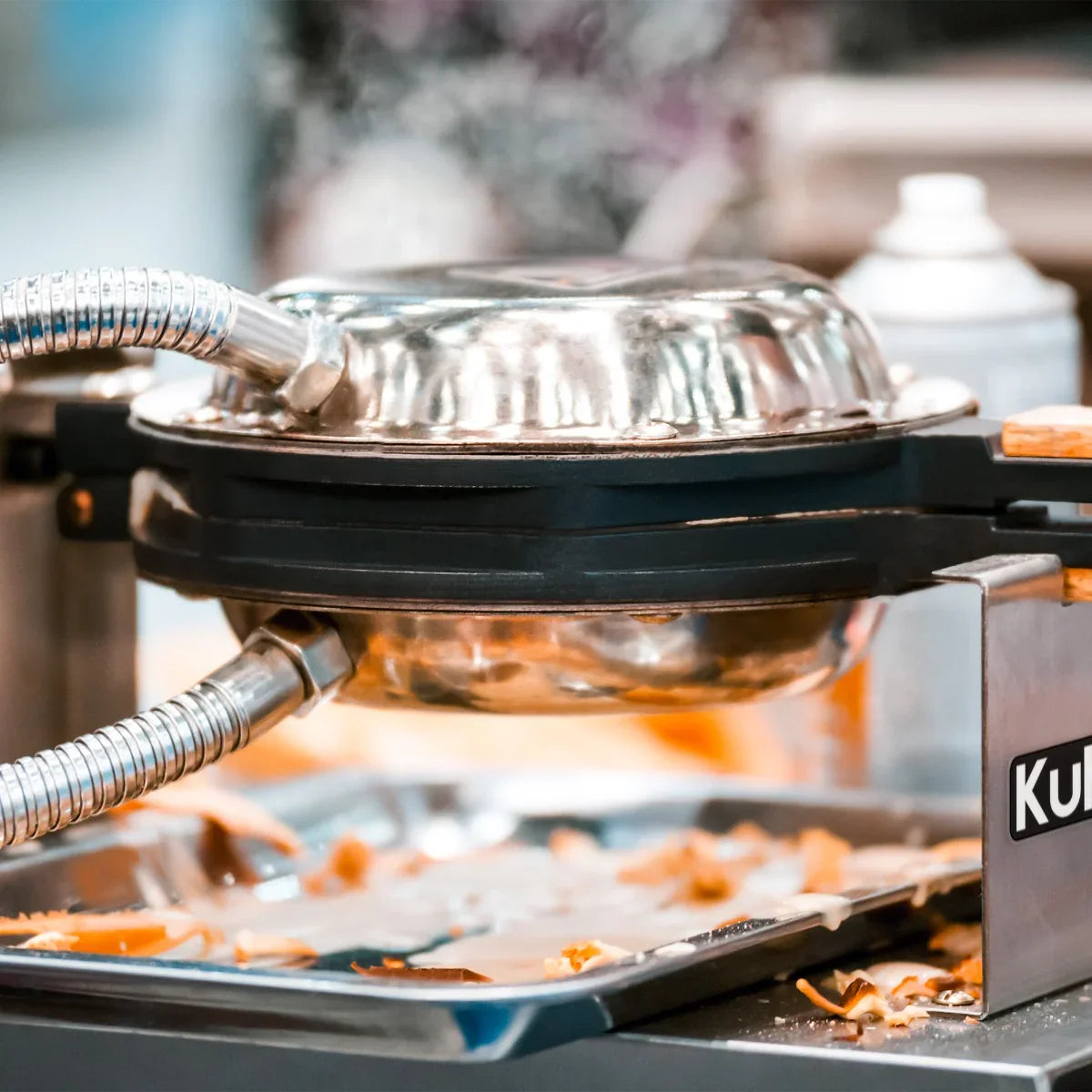 KuKoo Single Waffle Maker with stainless steel top, steam rising, and a tray beneath catching waffle drips and crumbs