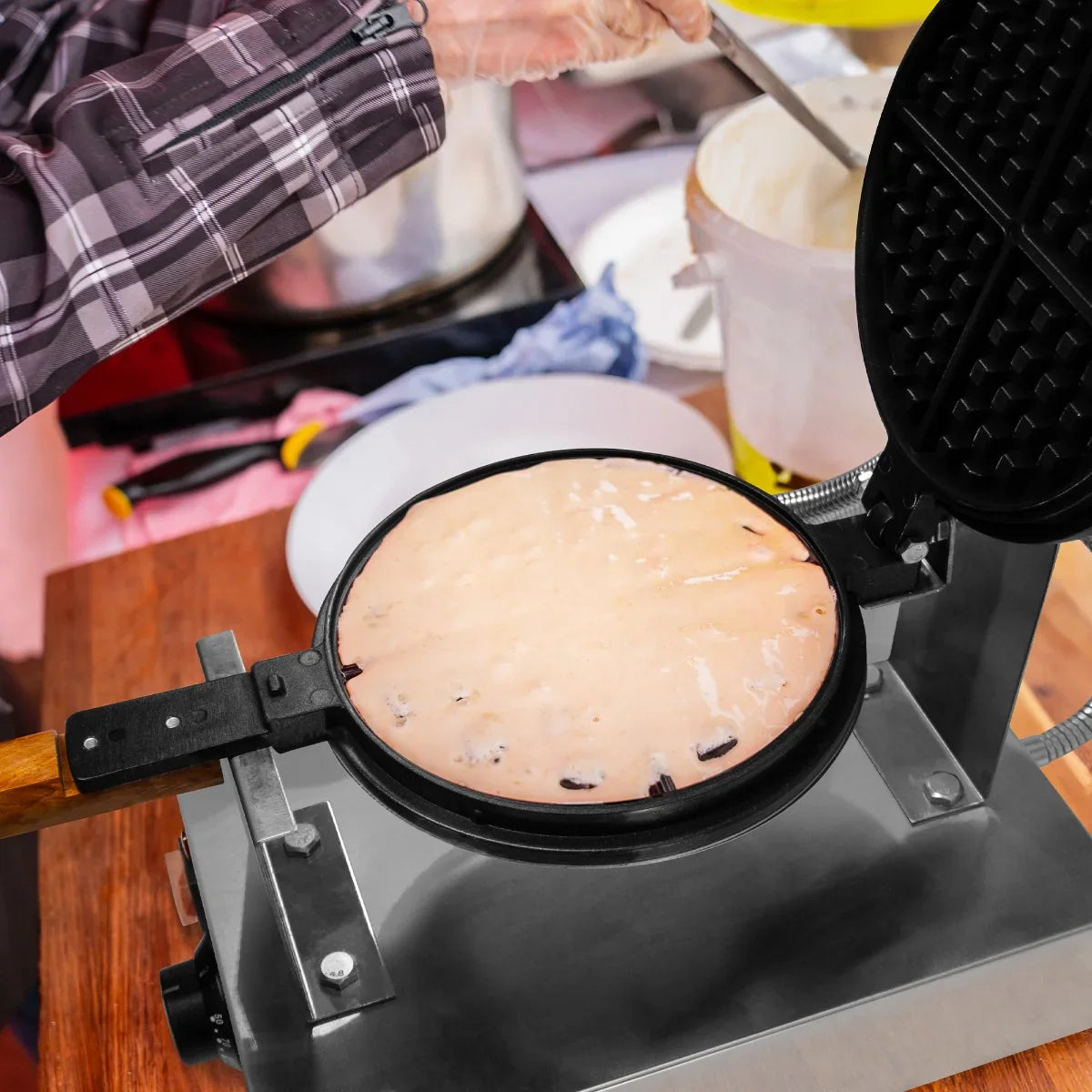 Single waffle maker in action, pouring batter into a round, non-stick cooking surface with a wooden handle, set on a stainless steel base