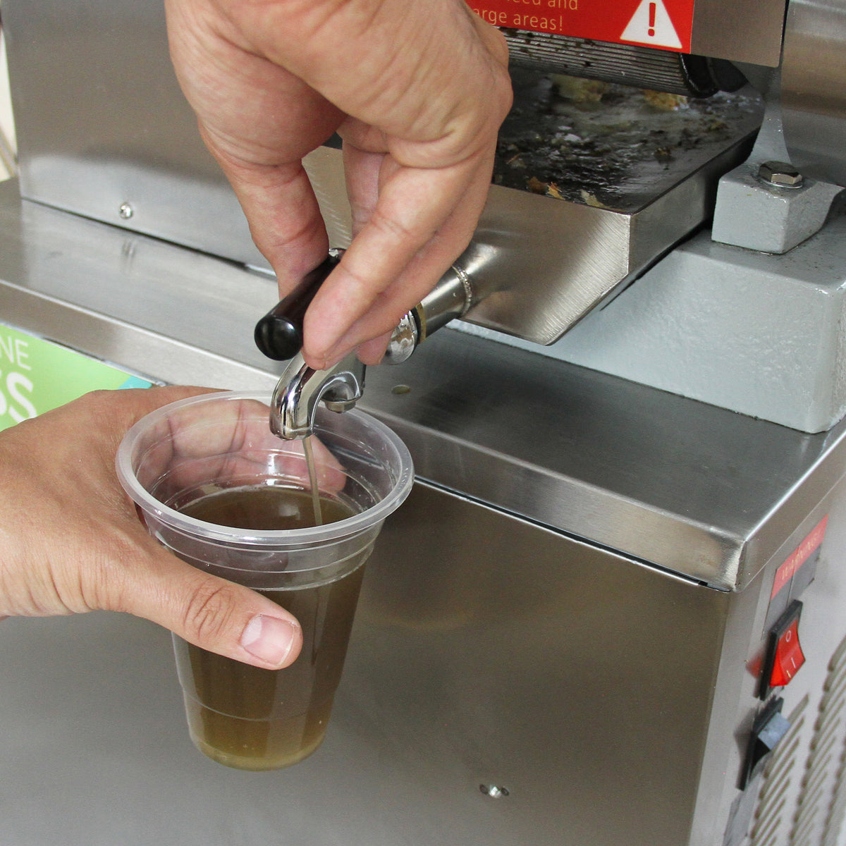 Hand pouring fresh sugar cane juice into a clear plastic cup from the KuKoo Sugar Cane Press, showcasing its metallic finish and sleek design