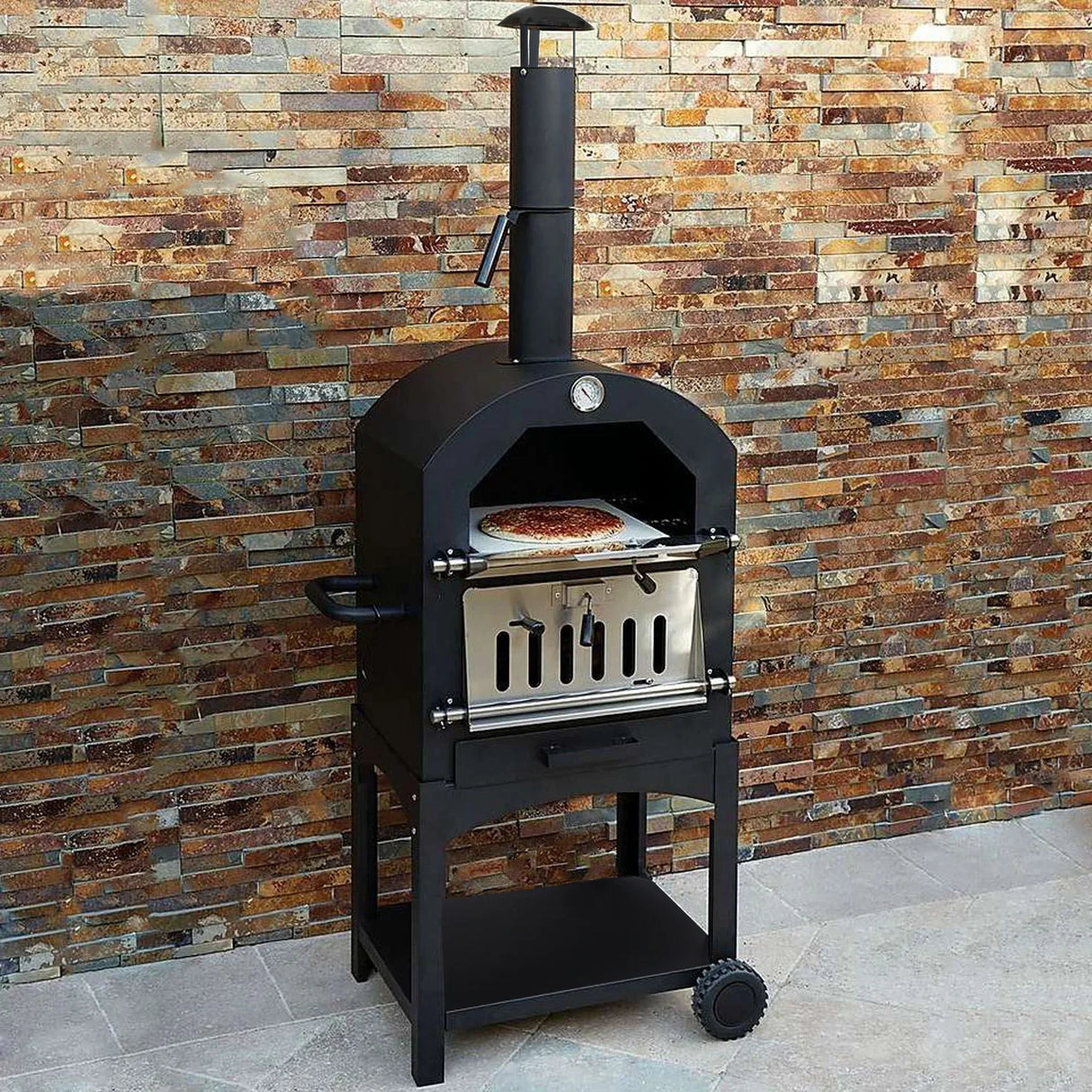 KuKoo outdoor pizza oven in black with stainless steel door, chimney, and a pizza on the stone baking surface against a stone wall