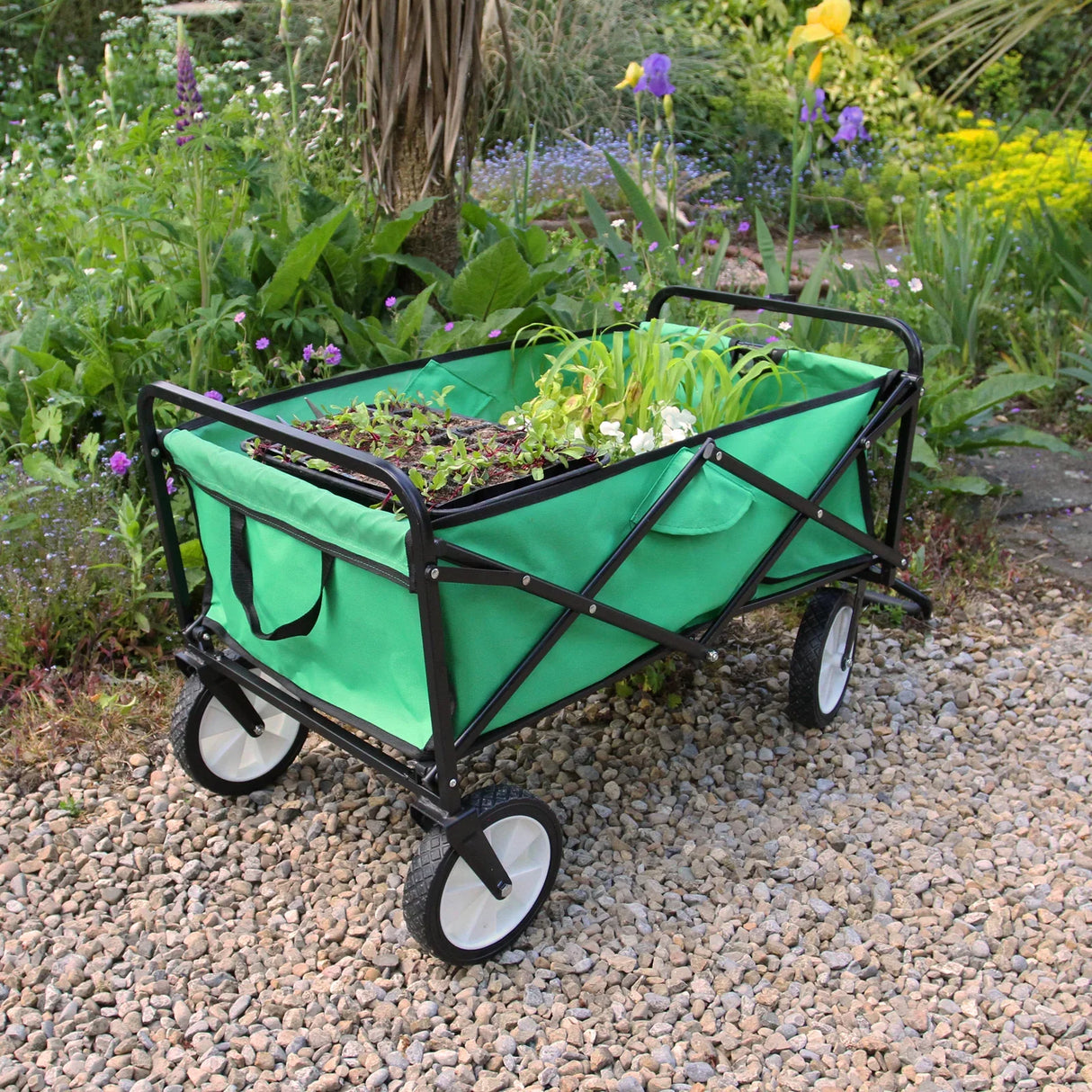 Foldable garden cart in vibrant green fabric with black frame, filled with plants, resting on gravel amidst colorful flowers