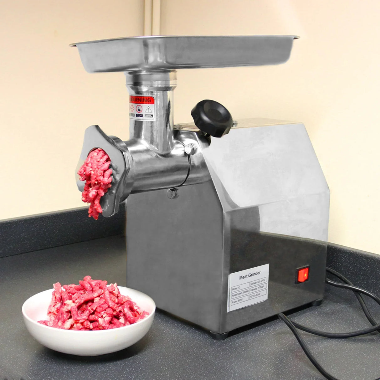 Stainless steel KuKoo Commercial Meat Grinder with a large feeding tray, freshly ground meat spilling into a white bowl on a counter