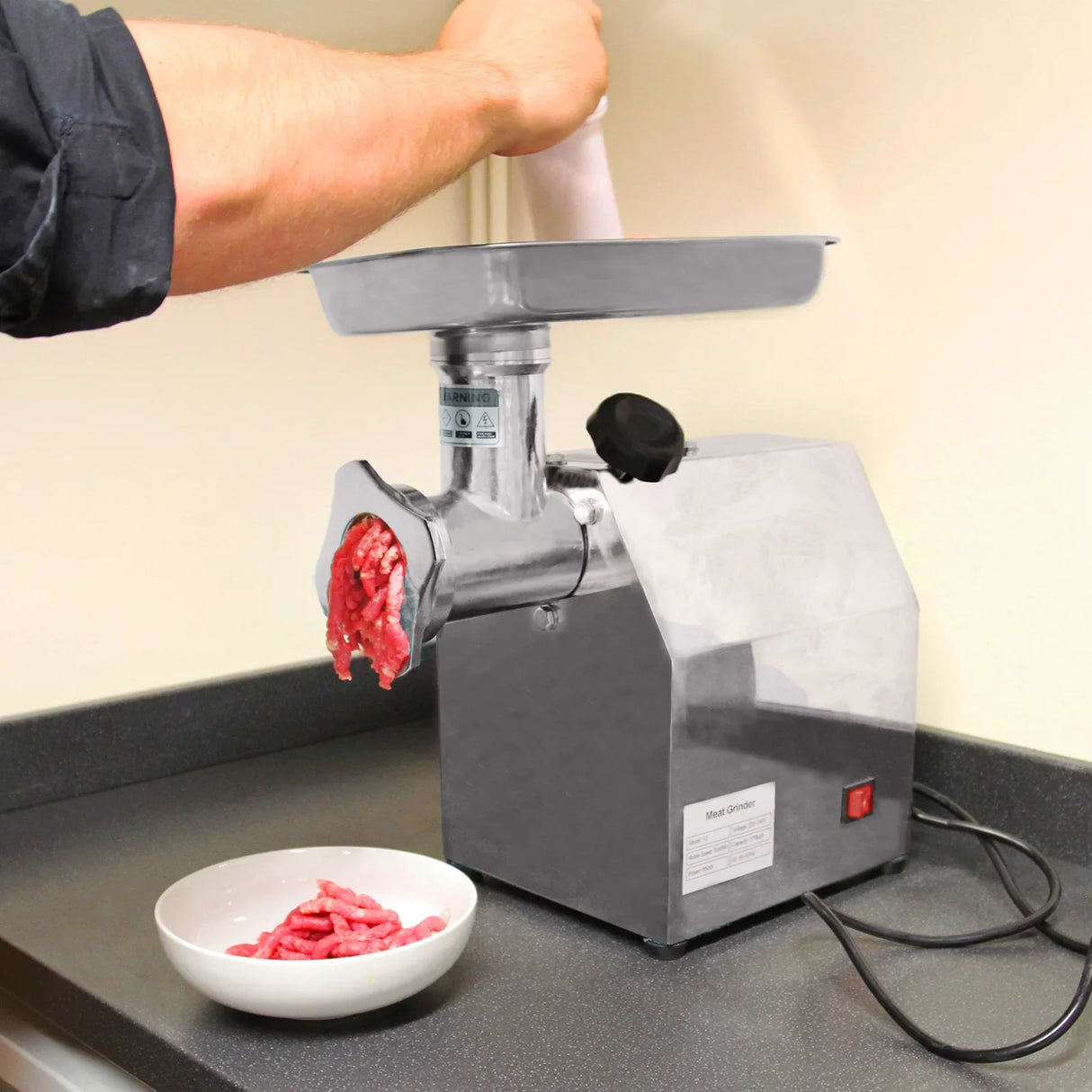 Stainless steel KuKoo Commercial Meat Grinder with ground meat being dispensed into a white bowl on a black countertop