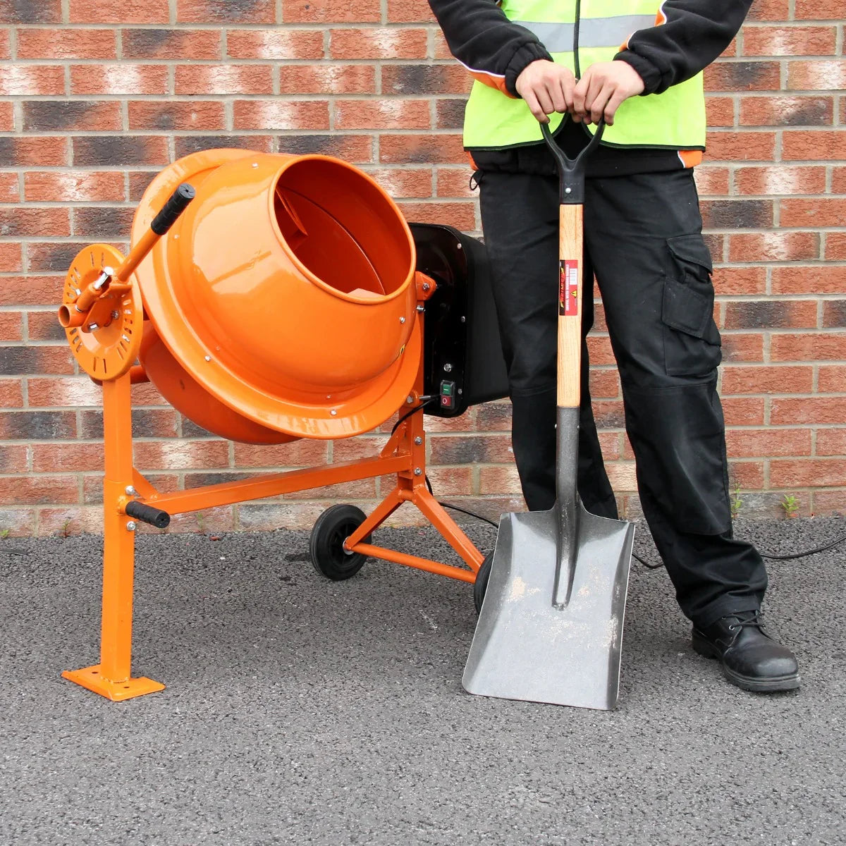 Cement mixer in bright orange with a sturdy metal frame, alongside a worker holding a shovel, set against a brick wall
