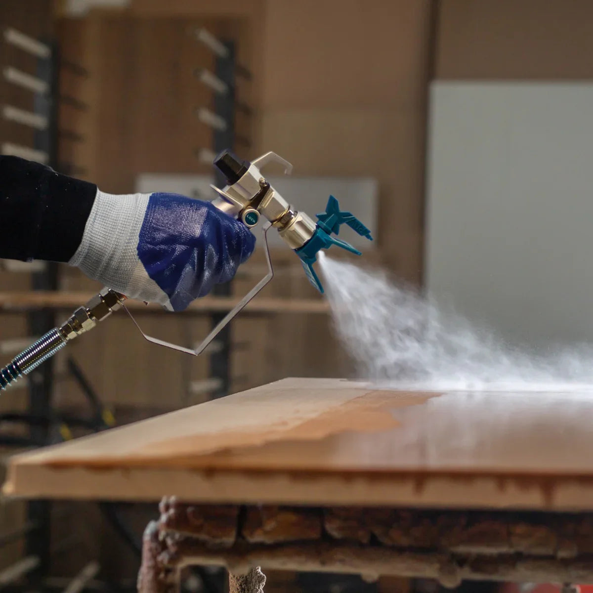 Maxblast Airless Paint Sprayer Gun in action, featuring a blue trigger, metallic body, and fine mist spraying over a wooden surface
