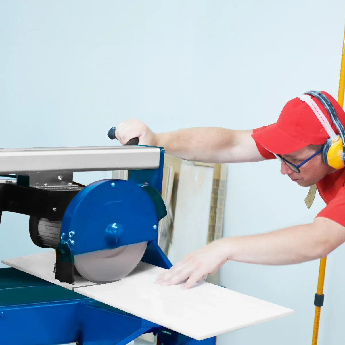 Wet tile cutter bench in blue with a 1200W motor, cutting a white tile, operated by a worker wearing safety gear and ear protection