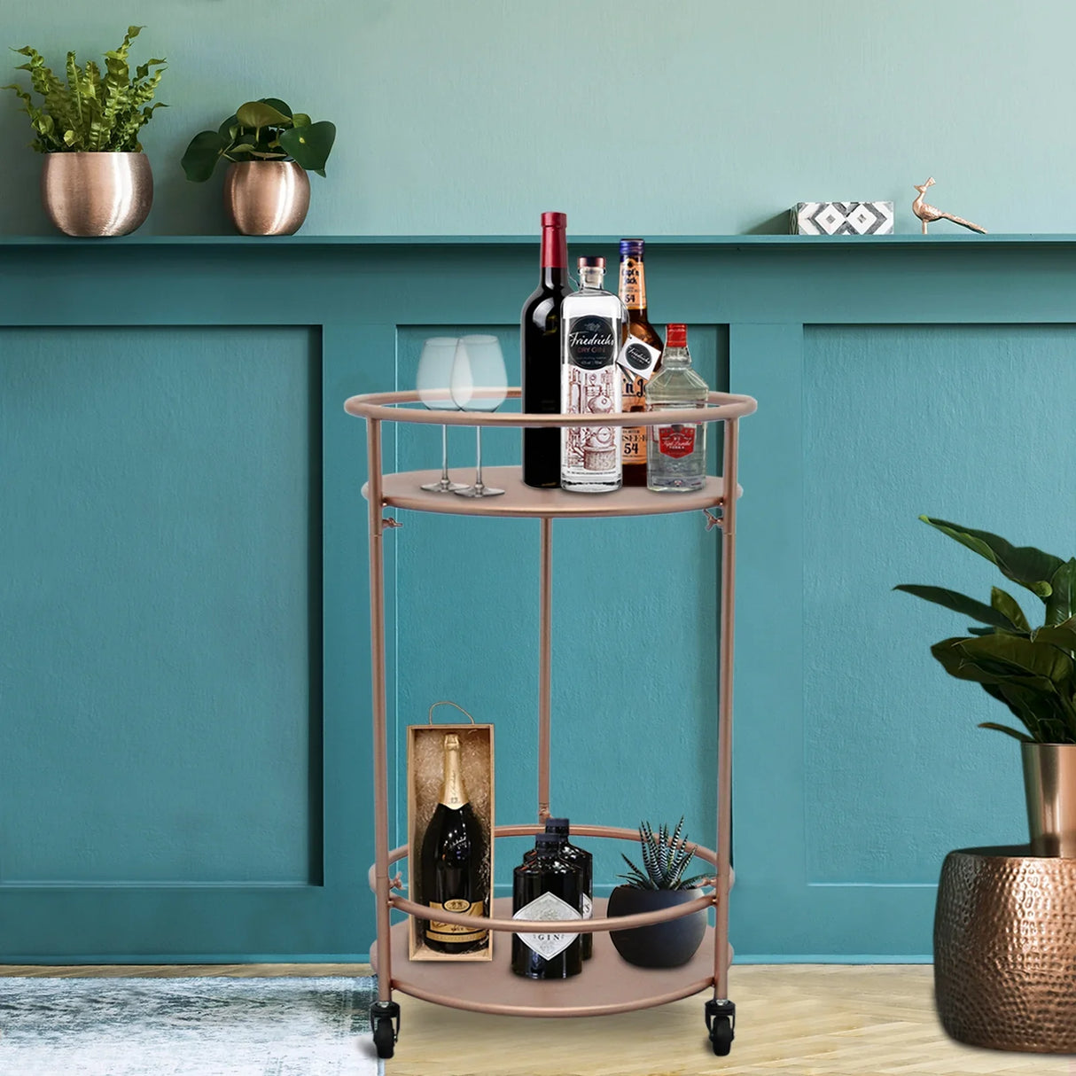 Rose gold small drinks trolley bar cart with two shelves, featuring glassware and assorted liquor bottles against a teal wall