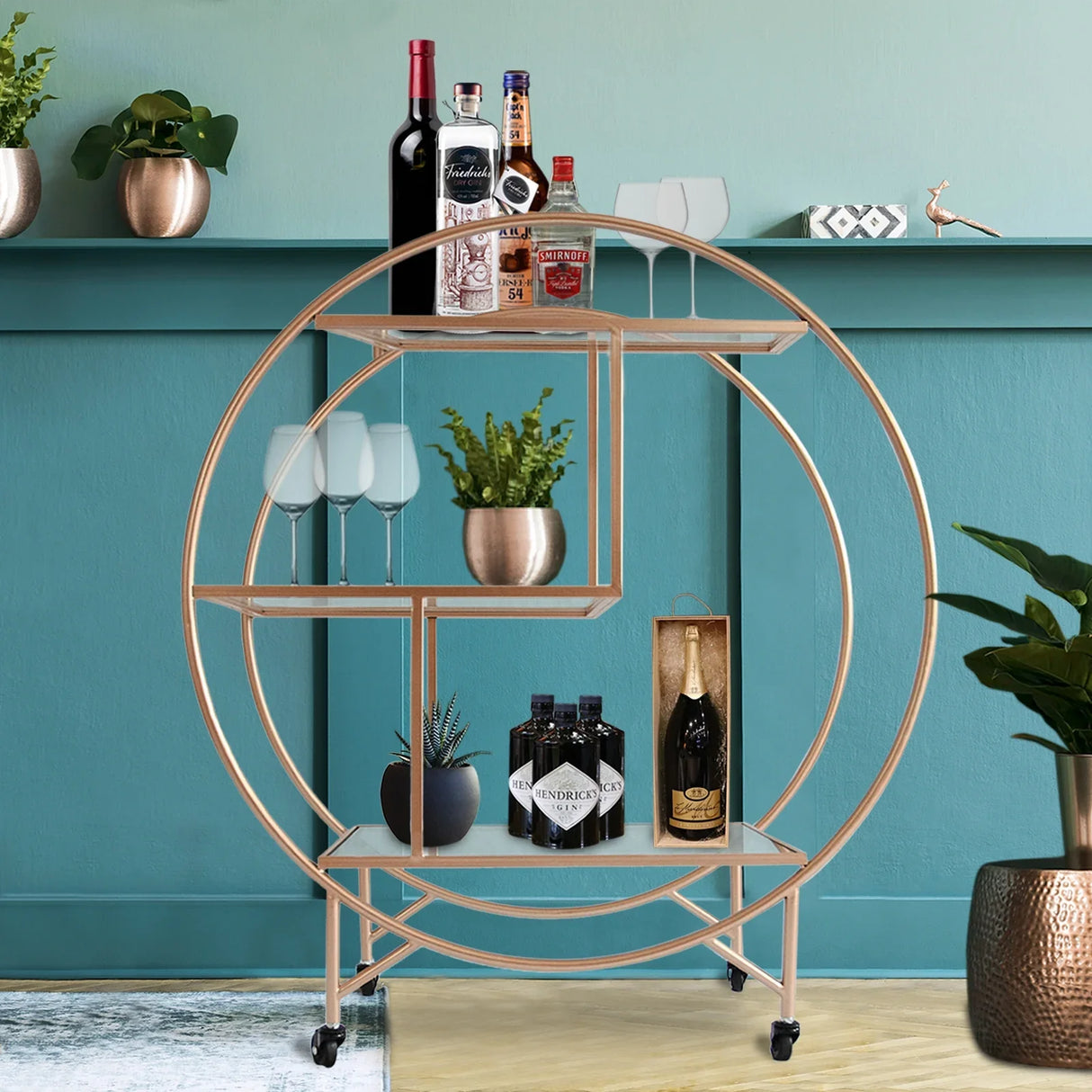 Large rose gold drinks trolley bar cart with circular design, glass shelves, wine bottles, and decorative plants against a teal wall