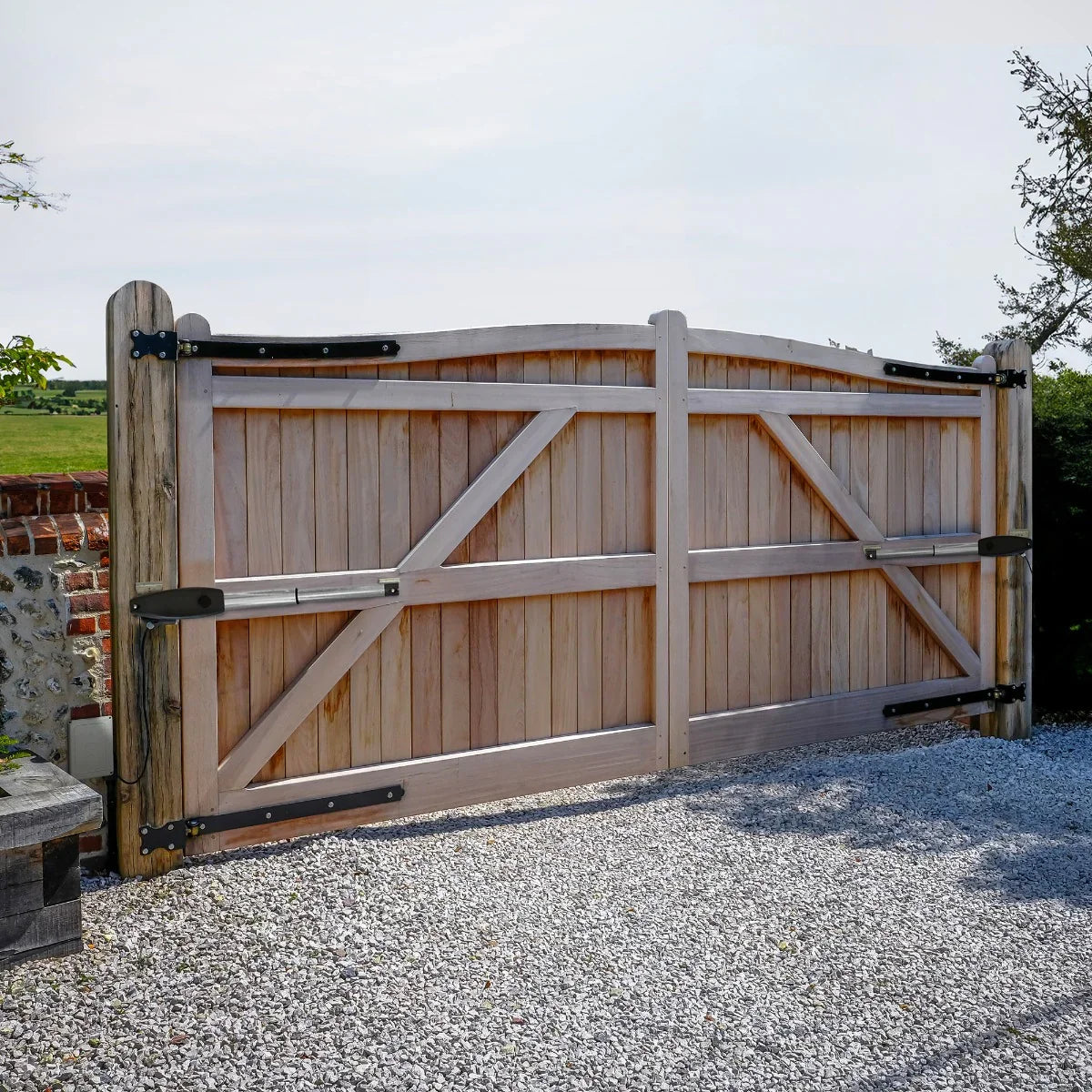Wooden electric swing gate with a curved top, featuring metal hinges and a smooth, natural finish, set against a gravel driveway