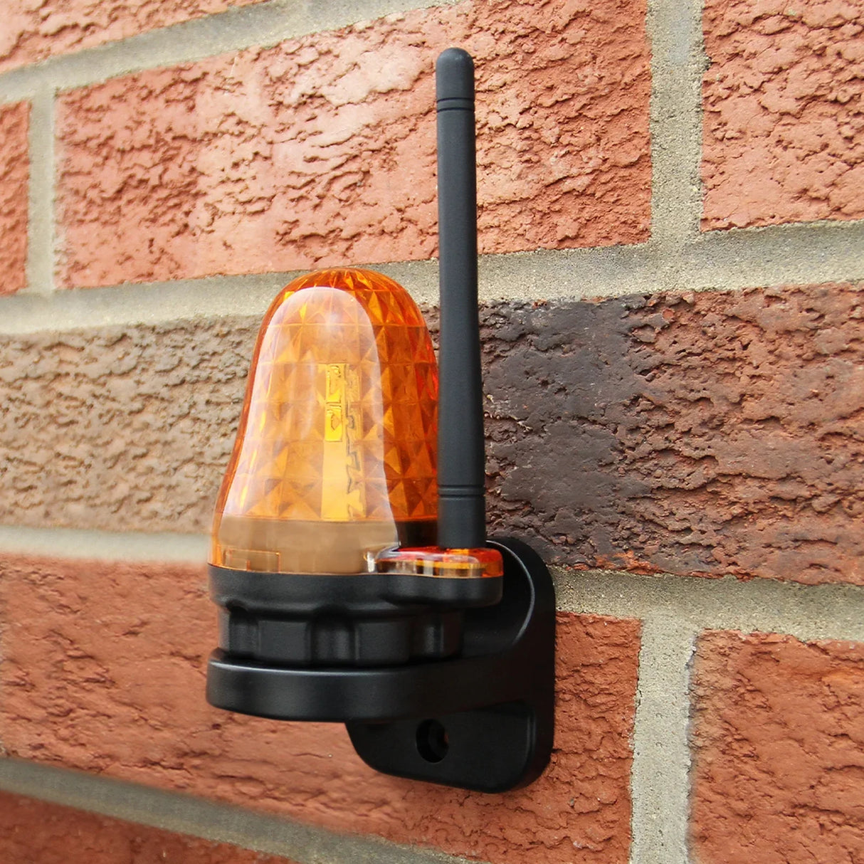 Bright orange warning light with a diamond pattern, mounted on a black base, featuring an antenna against a brick wall background